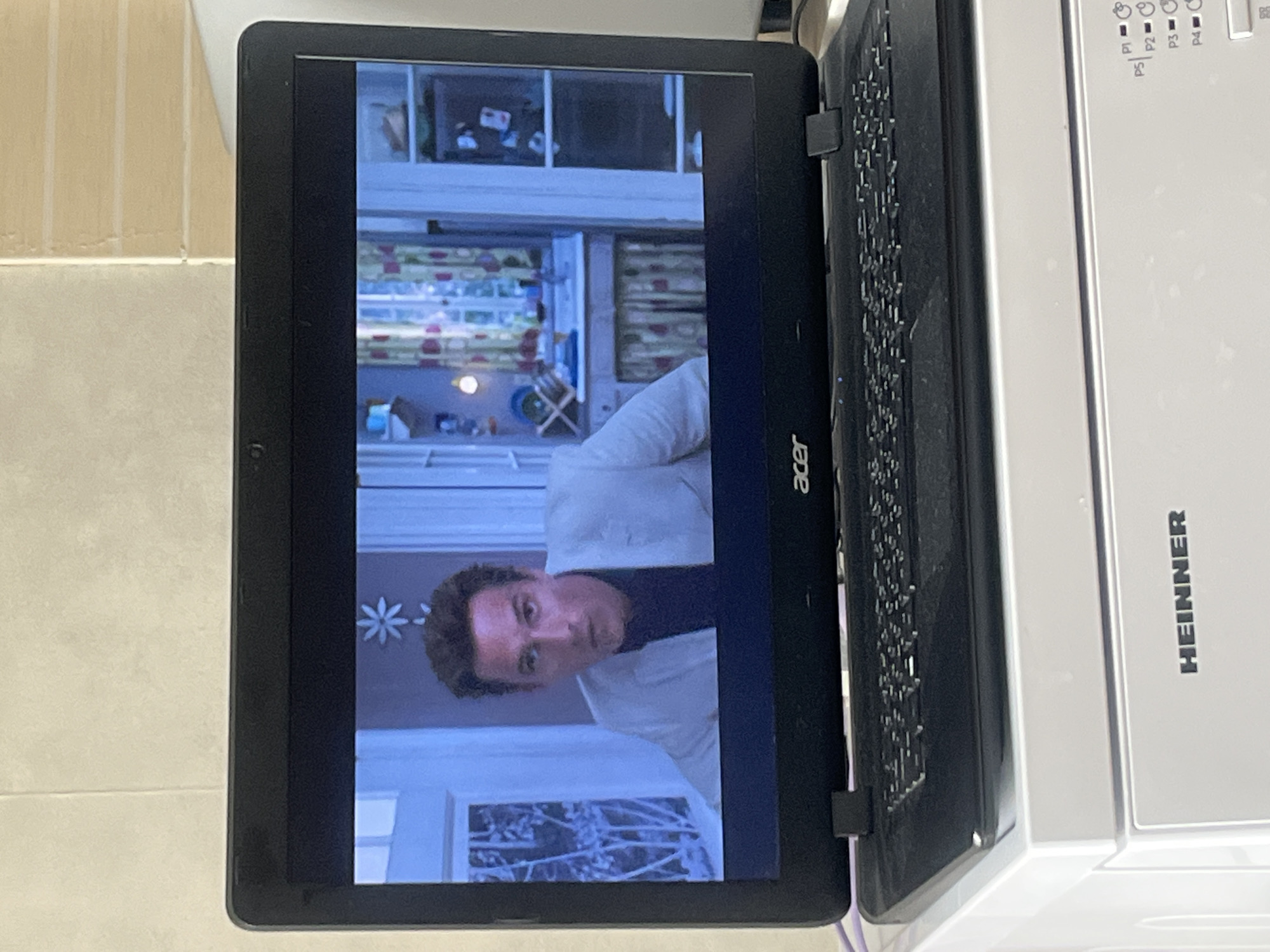 The image is a medium shot of a black Acer laptop placed on top of a white appliance, identified by the "HEINNER" logo. The laptop screen displays a scene from a movie, featuring a man with dark hair wearing a white suit jacket over a dark shirt. He is looking intensely towards the camera. The background on the screen depicts an interior setting, possibly a kitchen or a room with a window and curtains. The overall lighting suggests an indoor environment. The bottom portion of the image shows the keyboard of the laptop and the top of the white appliance with some indicator lights labeled P1 through P5. The overall scene is indoors, with a plain wall in the background above the laptop. There is no discernible context provided for the location in the city of Rybnitsa, Moldova.
