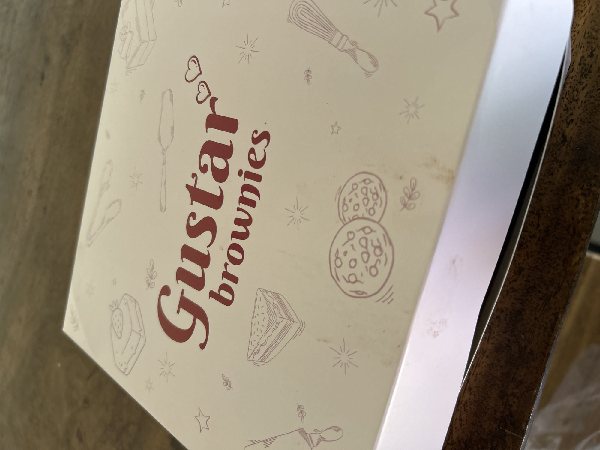 The image shows a close-up of a light beige box with a dark red, stylized logo and various line drawings of baked goods and baking tools. The logo reads "Gustar brownies" with two hearts above the "G." The drawings include a slice of cake with a strawberry, a tiered cake, a whisk, a rolling pin, and circular motifs that appear to be cookies or round pastries. There are also small decorative elements like stars and stylized leaves scattered across the box. The box is slightly open, revealing a white interior and a dark wooden surface beneath it, which suggests it might be on a table or counter. The lighting is natural and appears to be daytime. The overall impression is of a product packaging for a bakery or dessert brand. The text "Gustar brownies" is prominent on the box.