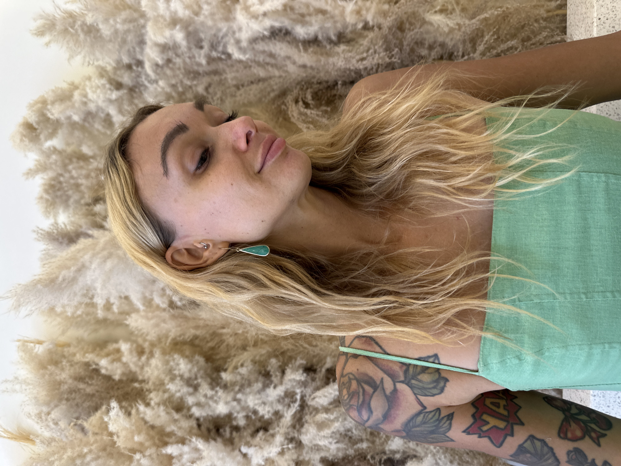 A woman with blonde hair is lying down in front of a backdrop of dried pampas grass. She is wearing a light green top. She has several tattoos on her right arm. She is wearing earrings. The image does not appear to be taken in Phu Thuy Ward, Vietnam, or depict anything about the location.