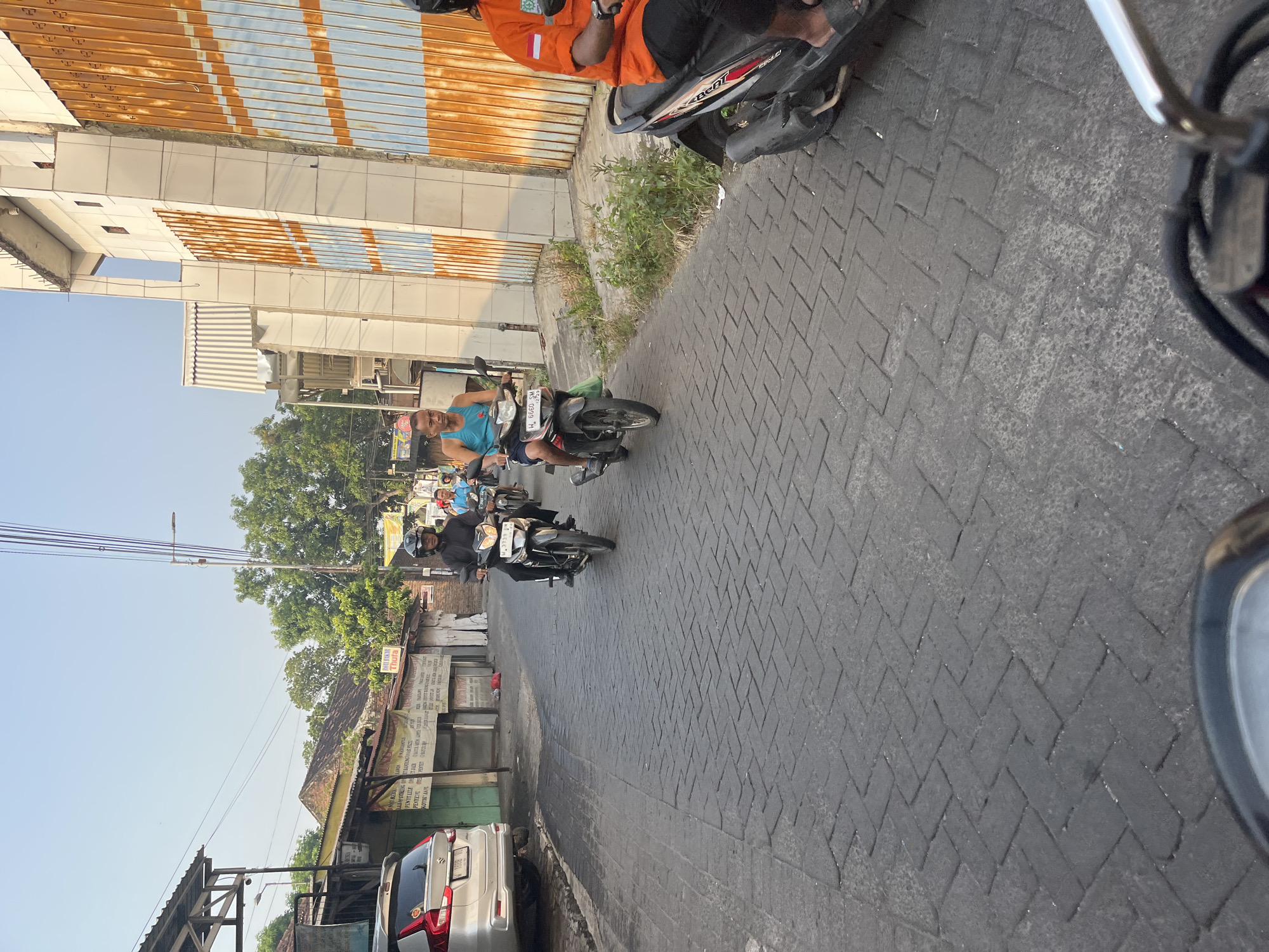 This image depicts a street scene in Tenggulunan, Indonesia. The road is paved with dark grey interlocking bricks laid in a herringbone pattern. On the left side of the road, a silver car is parked. Further down the street, a row of buildings with corrugated metal roofs and some signs are visible. The right side of the road is dominated by a large building with a white facade and a tall, orange and blue striped corrugated metal gate.
Several people are on motorcycles. In the foreground on the right, a person wearing an orange shirt is riding a black scooter. Further up the road, two men are riding motorcycles. One man in a blue tank top is riding a black motorcycle. Behind him, another man in a black jacket and helmet is also on a motorcycle. In the background, more motorcycles and people are visible. The sky is clear and pale blue, suggesting it is daytime, likely late afternoon due to the warm lighting. Green trees are present in the background, bordering the buildings and the street. The overall impression is of a busy, everyday street in an Indonesian town.