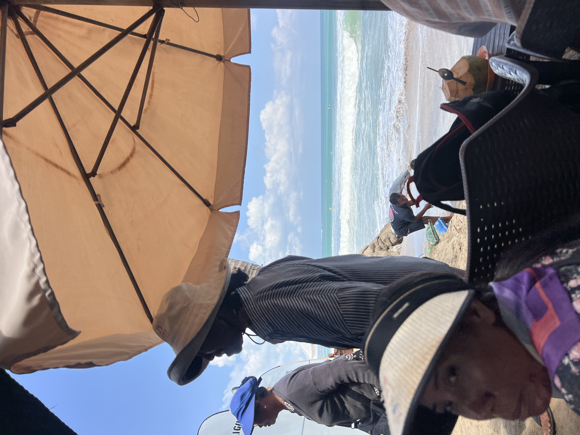 The image captures a scene at Kuta Beach, Indonesia, under a bright, partly cloudy sky. In the foreground, a woman wearing a visor and a purple and pink garment is partially visible, looking to her left. Behind her, an individual in a wide-brimmed hat and a striped shirt stands in profile, facing the ocean. To their left, another woman in a blue sun hat and dark clothing is also in profile, with a surfboard visible behind her.
The background features the turquoise ocean with moderate waves and several surfers in the water. A beach umbrella provides shade over the foreground elements. On the sandy shore, a person in a grey t-shirt is seen interacting with something on the sand. Various objects, including chairs, a backpack, and a coconut drink with a spoon, are present in the immediate foreground, suggesting a relaxed beachside setting. The overall atmosphere is one of a casual day at the beach.