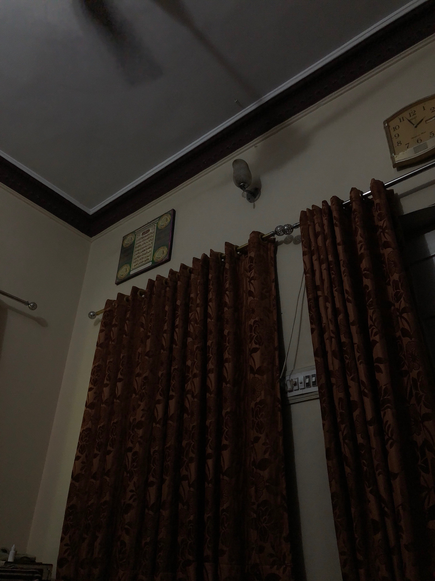 The image depicts an indoor scene, likely a room, with elements suggesting a living space. The most prominent features are the dark brown, patterned curtains hanging from a curtain rod that spans across the frame. The curtains have a floral or leaf-like motif. Above the curtains, a portion of a cream-colored ceiling is visible, with dark decorative trim along the edge where it meets the wall. A faint shadow of a ceiling fan is visible on the ceiling.
On the wall, to the right, a vintage-style analog clock with a light-colored face and dark numbers is mounted. The time appears to be approximately 12:35. To the left of the clock, a small wall-mounted light fixture is visible. Further to the left, a framed artwork or religious text with Arabic calligraphy is displayed on the wall. There are also horizontal metal rods with spherical finials, possibly curtain rods or decorative elements, extending from the walls.
In the lower section of the image, on the right side, a row of light switches or electrical outlets is partially visible. The overall lighting is dim, creating shadows and highlighting the textures of the curtains and walls. The scene does not contain any people. Given the context of Larkana, Pakistan, this scene could represent a room in a residential house or a hotel, common in cities like Larkana, which is a significant city in Sindh province.