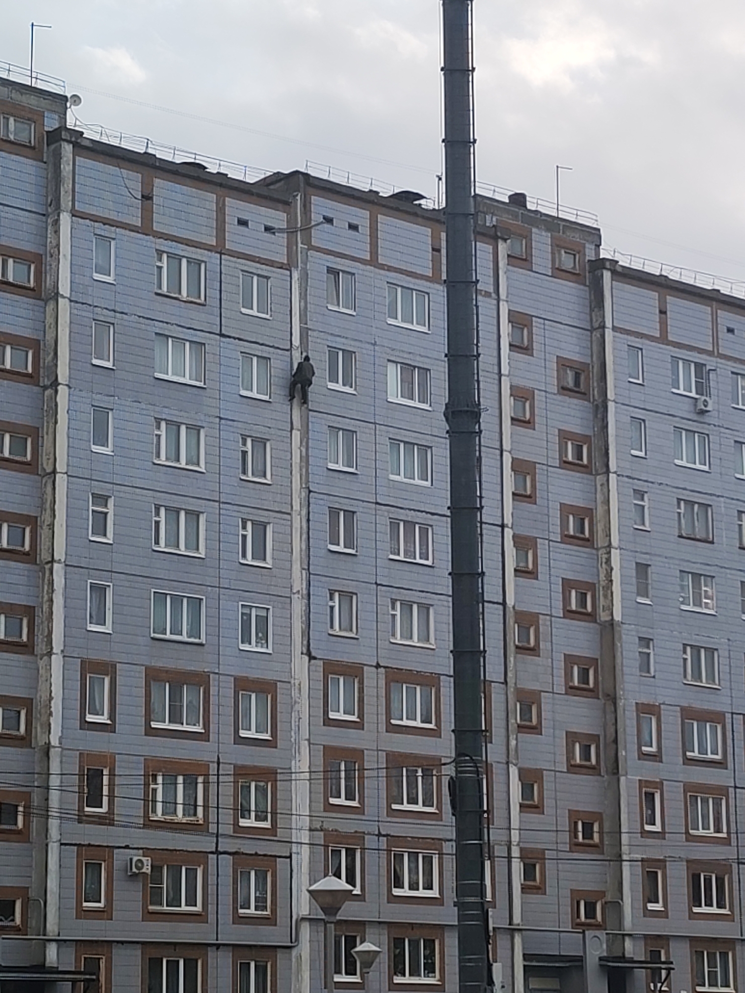 The image depicts the facade of a multi-story residential building in Izhevsk, Russia. The building is constructed of prefabricated concrete panels, arranged in a grid pattern. A person is climbing a section of the building's facade. The architecture is typical of Soviet-era urban construction. A tall, cylindrical pipe runs vertically along the exterior of the building. The sky is overcast.