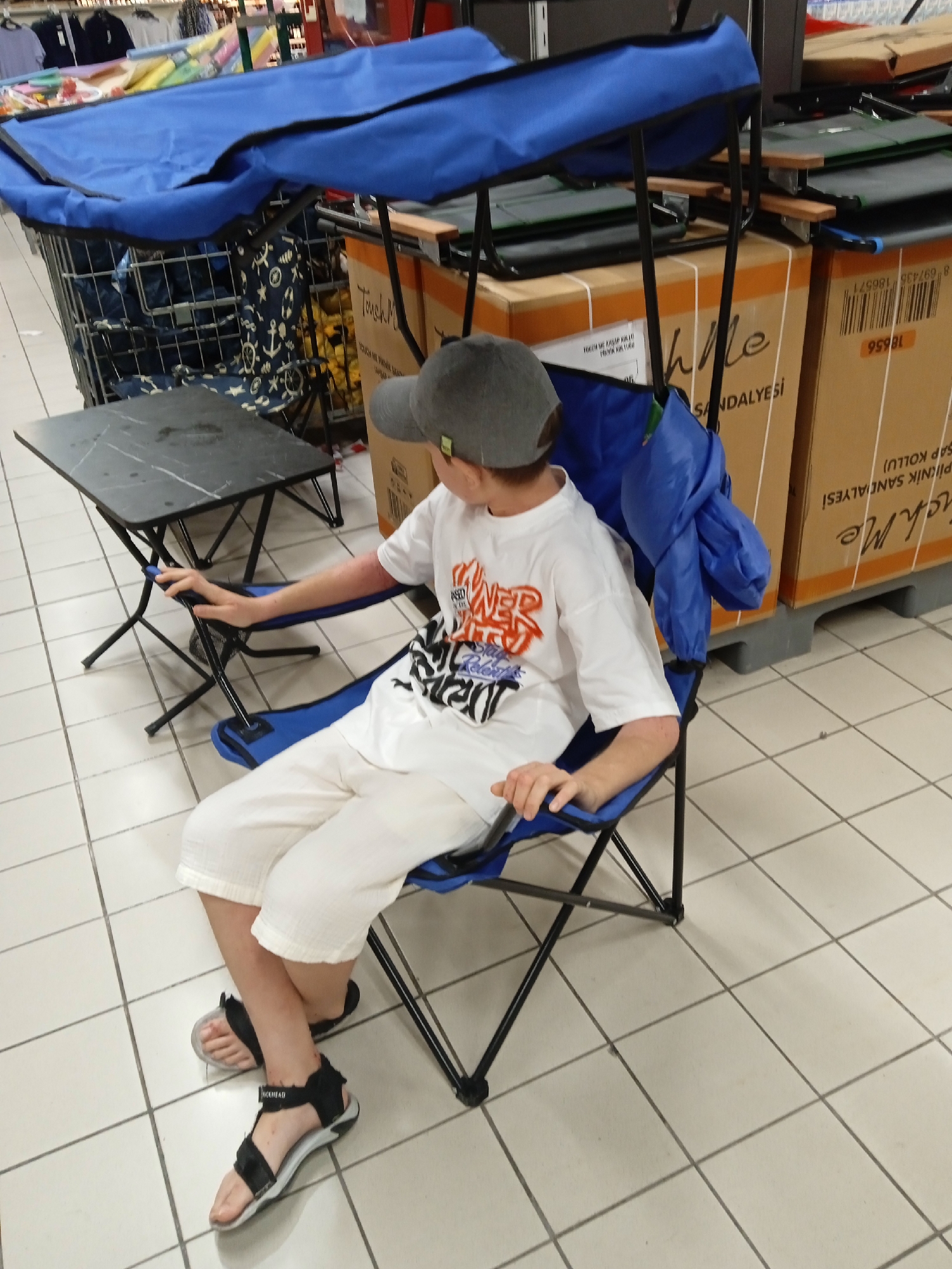 A young person wearing a grey baseball cap, a white t-shirt with orange and black graphics, and white shorts sits in a blue folding chair in a retail store in Yenişehir, Turkey. The person is also wearing black and grey sandals. A small black folding table with a marbled top is positioned to their left. The chair and table are set up in front of a shopping cart. In the background, there are stacks of cardboard boxes, some with product information and barcodes. The floor is tiled in a grid pattern. The lighting suggests an indoor, commercial environment.