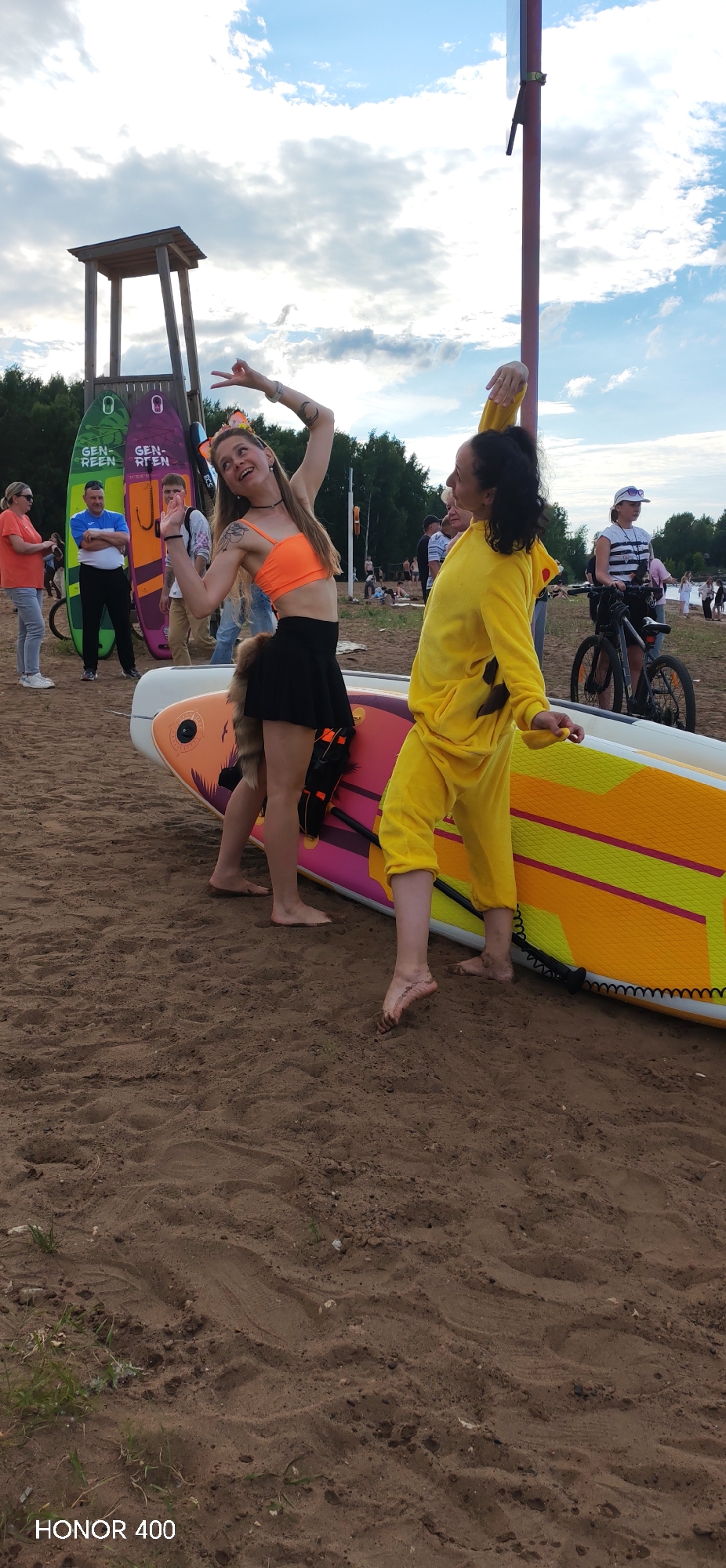 Two young women are posing in front of a paddleboard on a sandy beach. The woman on the left is wearing an orange crop top and a black skirt, with what appears to be a fox tail attached to her back. She has her arms raised in a playful pose and is smiling. The woman on the right is wearing a yellow Pikachu costume. They are both barefoot.
In the background, several paddleboards are standing upright. Behind them, a wooden lifeguard tower is visible, along with trees and a cloudy sky. Several other people are scattered around the beach, some are walking, and others are near bicycles. One person is riding a bicycle in the middle-ground.
The scene appears to be a casual outdoor gathering or event, possibly related to water sports given the paddleboards. The lighting suggests it is daytime, likely afternoon, with some cloud cover. The overall atmosphere seems relaxed and festive, with the two women clearly enjoying themselves.
The visible text at the bottom of the image reads "HONOR 400".