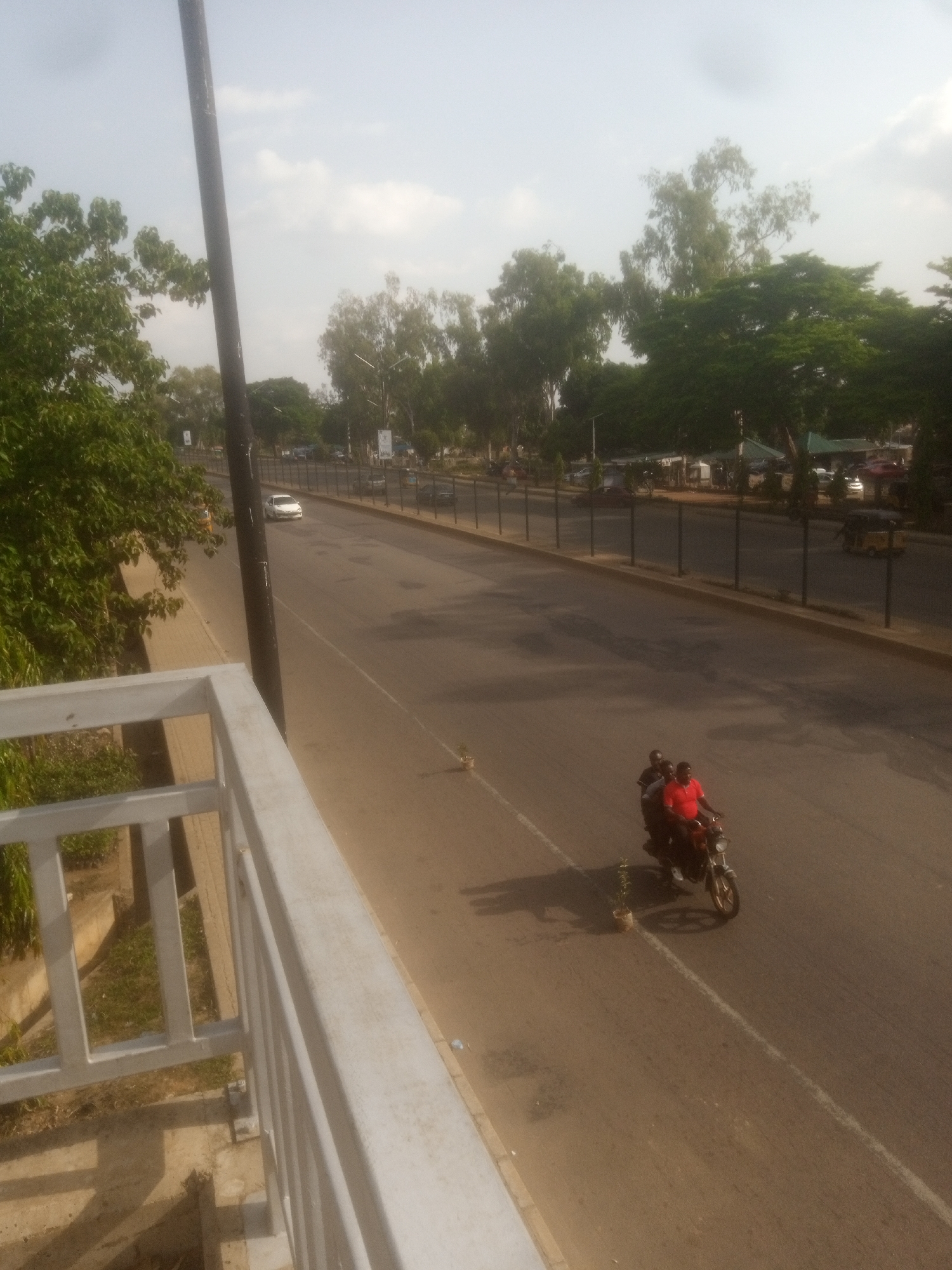 The image shows a street scene in Unguwan Dosa, Nigeria. The foreground features a white metal railing, suggesting a raised viewpoint, possibly a bridge or an elevated walkway. Beyond the railing, a paved road stretches into the distance, lined with trees and a dark fence. The sky is overcast, indicating it could be morning or late afternoon, with soft, diffused light.
The main activity visible is on the road where two people are riding a motorcycle. The rider, wearing a red shirt, and a passenger are moving away from the viewer. Their presence suggests everyday transportation. In the background, a white car is driving on the road, and further down, a yellow three-wheeled vehicle, possibly a tuk-tuk, is visible. There are also buildings and trees on the other side of the road, hinting at an urban or semi-urban environment. A small potted plant sits on the side of the road near the motorcycle, a small detail adding to the scene's ordinariness. No visible text is present in the image. The overall impression is a calm, everyday street scene in a populated area.