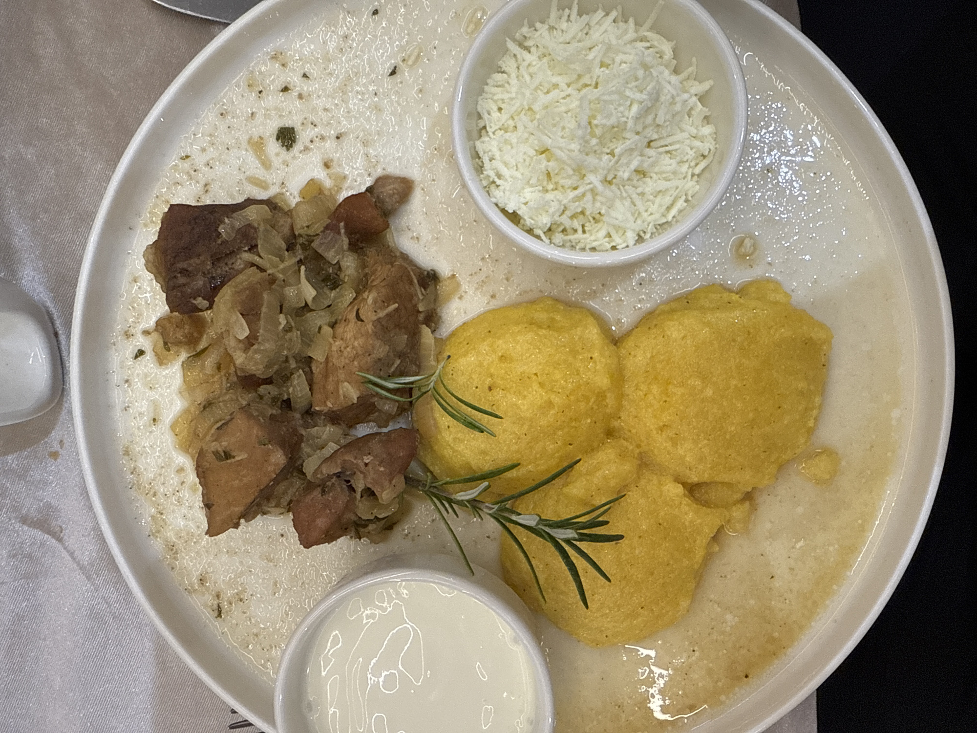 The image shows a plate of food on a white tablecloth. The plate is white and round, with a slight rim. The food consists of several pieces of browned meat mixed with onions, three portions of yellow polenta, a small bowl of white cream or yogurt, and a small bowl of grated white cheese. A sprig of rosemary is placed on top of the polenta. The background is a blurred white tablecloth. The image appears to be a close-up of a meal, likely taken indoors. Based on the food presentation and style, this is likely a traditional Moldovan dish. Băcioi is a commune in Moldova, known for its agricultural products and traditional cuisine. This meal is consistent with what might be served in a restaurant or home in this region of Moldova.