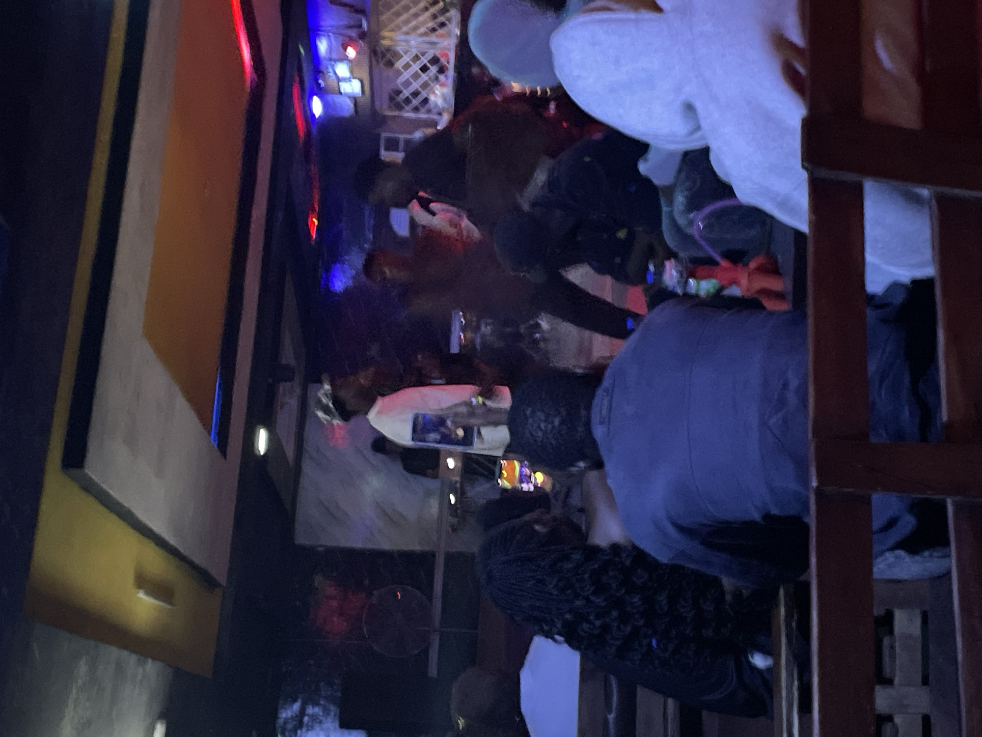 The image depicts an indoor scene, likely a bar or club, with patrons seated and standing. The lighting is dim and atmospheric, with red and blue neon lights visible in the background, suggesting a nighttime setting. A person in the background appears to be performing or speaking on a stage, possibly a musician or DJ, indicated by the presence of equipment. Patrons are casually dressed, with some engaged in conversation. The overall impression is one of a social gathering or entertainment venue. No specific geographical markers for Durimi, Nigeria are present in the image.