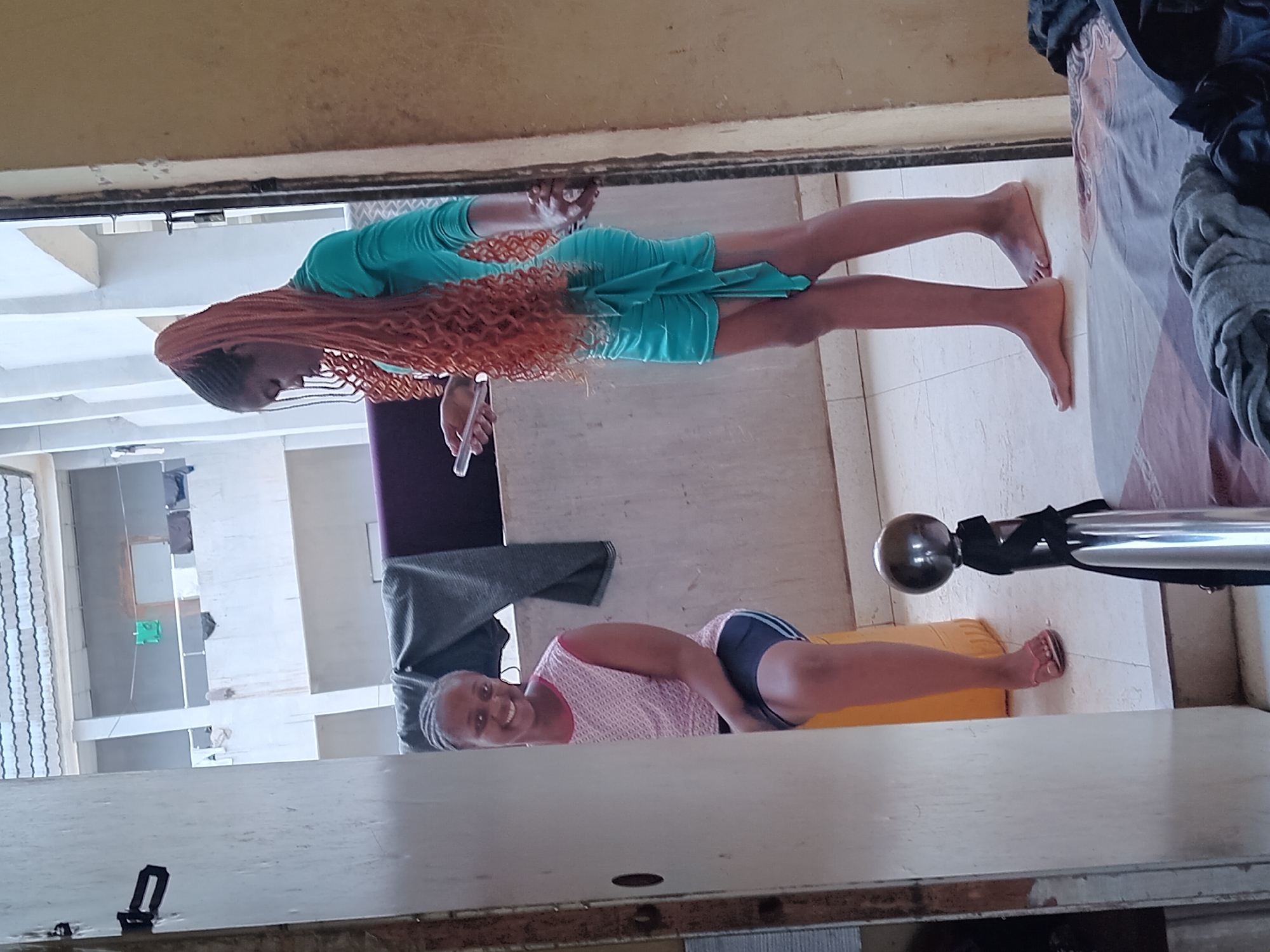 Two women are present in an indoor setting. One woman is wearing a teal dress and has long, reddish-brown hair. She is holding a cellphone. The other woman is wearing a pink tank top and dark shorts, and she is smiling. The environment includes a bed with grey bedding, a purple object, and an exterior view of a building, possibly in the city of Umueshi, Nigeria. The specific actions and interactions between the women are not explicitly evident.