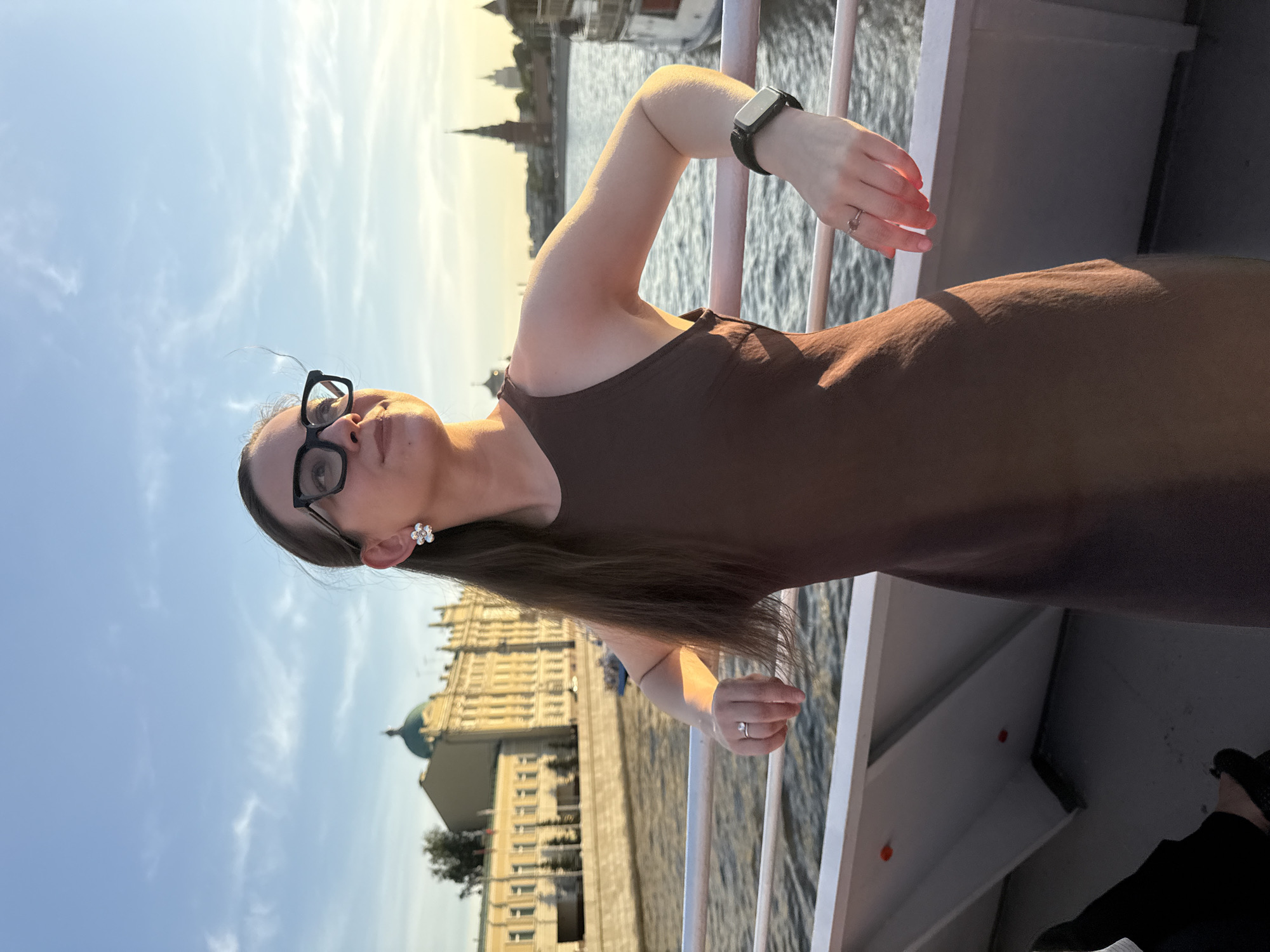 A woman is standing on a boat, presumably touring the Moscow River in Moscow, Russia. She is wearing glasses, a dark dress, and a smartwatch. She has long brown hair. Her hands are resting on the boat's railing, and she is wearing a ring on each hand. In the background, there are historical buildings and a dome-shaped structure. The sky is bright with a few clouds, suggesting it is either early morning or late afternoon. The Moscow Kremlin complex is visible on the background.