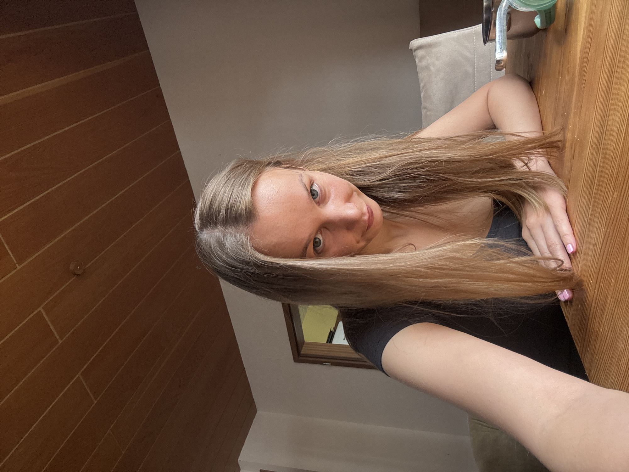 A young woman with long, blonde hair is leaning on a wooden table. She is wearing a black t-shirt. Her right hand is resting on the table, and her left arm is bent at the elbow. She is looking at the camera with a neutral expression. The background shows a wooden wall, a neutral-colored wall, and a glimpse of a window. There is a faucet and a green object on the table behind her. The overall lighting suggests it is daytime. The setting appears to be indoors, possibly a home or a cafe, based on the furniture and decor. The image is taken from a high angle, looking down at the woman.