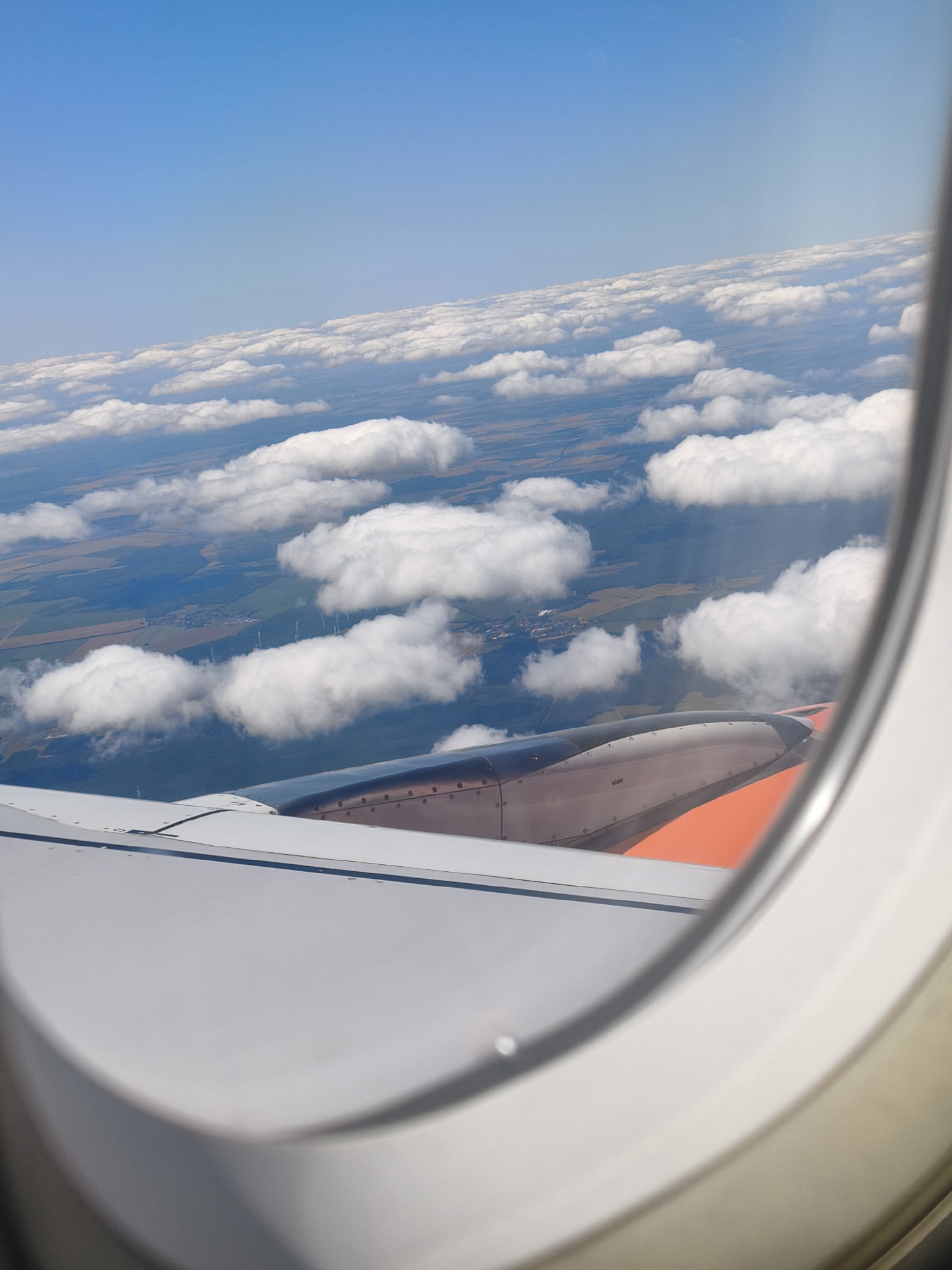 The image, taken from an airplane window, offers a view of the landscape below and the aircraft's wing. The sky is a clear, bright blue, with scattered white clouds creating a textured layer between the plane and the earth.
Below the clouds, the land appears as a patchwork of agricultural fields and green forested areas. In the distance, a cluster of wind turbines is visible, indicating a rural or semi-rural setting. A small village or settlement can be discerned amidst the fields, with houses and structures suggesting human habitation.
The aircraft's wing, a significant portion of the foreground, is predominantly white and grey. The engine housing, with its metallic and orange accents, is partially visible, extending from the wing. The window frame, a thick white border, encloses the view.
Based on the visible wind turbines and the landscape, this scene is likely located in the vicinity of Baruth/Mark, Germany, a region known for its agricultural land and the presence of wind farms.