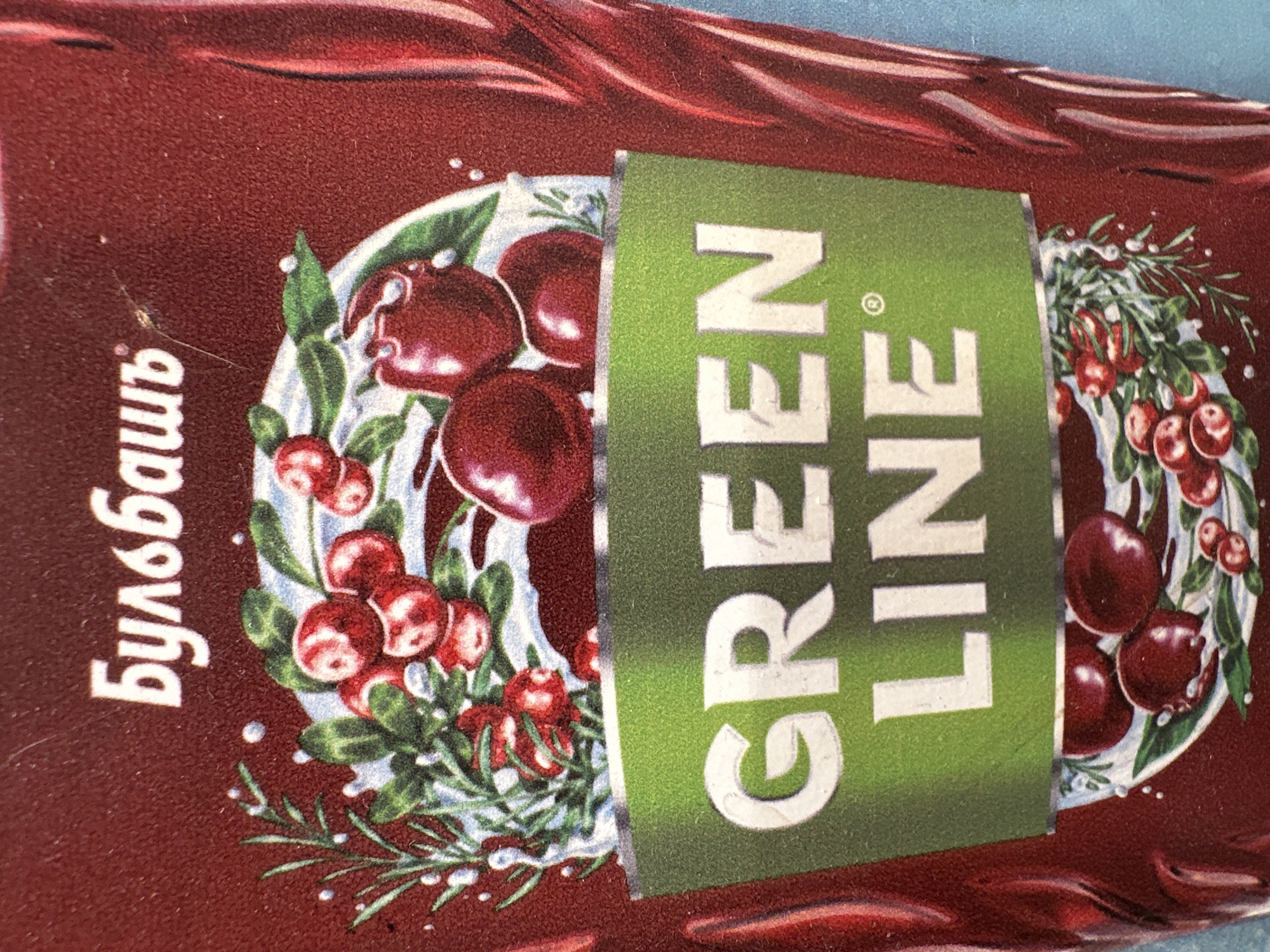 The image is a close-up of a product package, likely a beverage or food item, with a dark red background. The main subjects are several sprigs of cranberries with green leaves, depicted as if splashing or being poured. The cranberries are a deep red, round, and glossy, with some appearing to be in clusters. White splashes and droplets are scattered around the cranberries and leaves, suggesting liquid or a refreshing quality.
In the center of the image, a green rectangular label is placed diagonally. It contains white text that reads "GREEN LINE". Below this, a section of Cyrillic text is visible vertically on the left side of the package, reading "бульбашь".
The overall impression is one of freshness and natural ingredients, with the vibrant red of the cranberries contrasting with the green of the leaves and the label. The setting is not explicitly depicted, but the product packaging suggests it is a commercial item. The lighting appears to be bright and even, highlighting the details of the product design. There are no people or any specific time of day or weather conditions indicated. The location context is Minsk, Belarus.