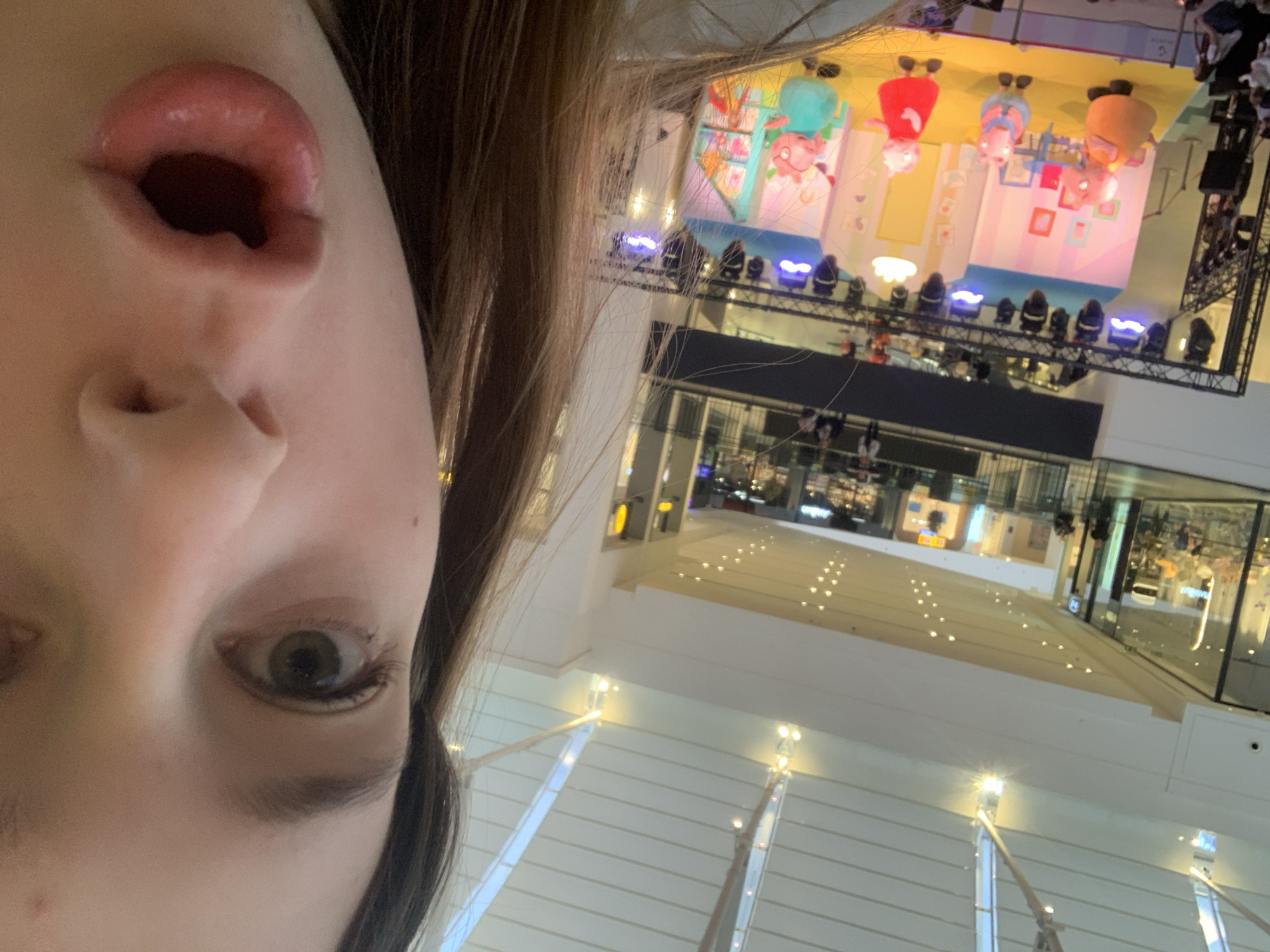 A close-up, diagonally oriented selfie captures a person's right eye and slightly parted, glossy lips in the foreground. The person has fair skin and light brown hair partially visible on the right side of the frame. Their eye looks directly at the camera.
The background, rotated 90 degrees clockwise relative to the person's face, depicts a brightly lit indoor commercial or event space, likely a shopping mall in Belgrade, Serbia. An elevated stage or display platform dominates the upper background. On this platform, large, colorful figures resembling the Peppa Pig family (Peppa, George, Mummy Pig, and Daddy Pig) are prominently displayed, standing within themed room-like sections (yellow, blue, pink) decorated with small frames. A row of bright spotlights illuminates the characters from below.
Further into the background, multiple levels of the building are visible, characterized by polished floors, glass railings, and numerous artificial lights, suggesting shops or public areas within a modern indoor complex. The scene captures a lively atmosphere, possibly during a children's event or promotional display. No discernible text is visible.