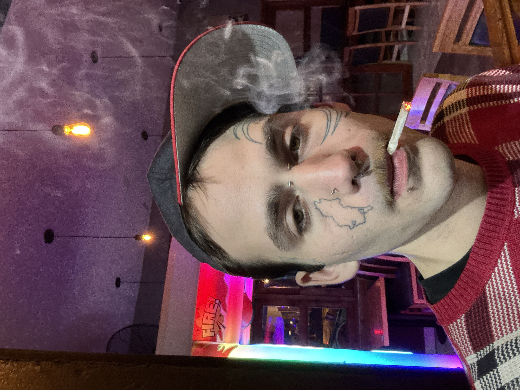 A close-up, tilted view of a man smoking a cigarette, likely in a bar setting. He has a gray baseball cap with a red brim, and a red and white patterned sweater. Distinct tattoos adorn his face, including curved lines above his eyebrows and a map-like shape near his cheek. He has multiple piercings on his face. Smoke is visible emanating from the cigarette. The background reveals a dimly lit bar with various lighting elements, including hanging Edison-style bulbs and illuminated signs. A glass with the word "FIRE!" on it is visible. The location appears to be indoors. There is no indication that this image is taken in Buenos Aires, Argentina.