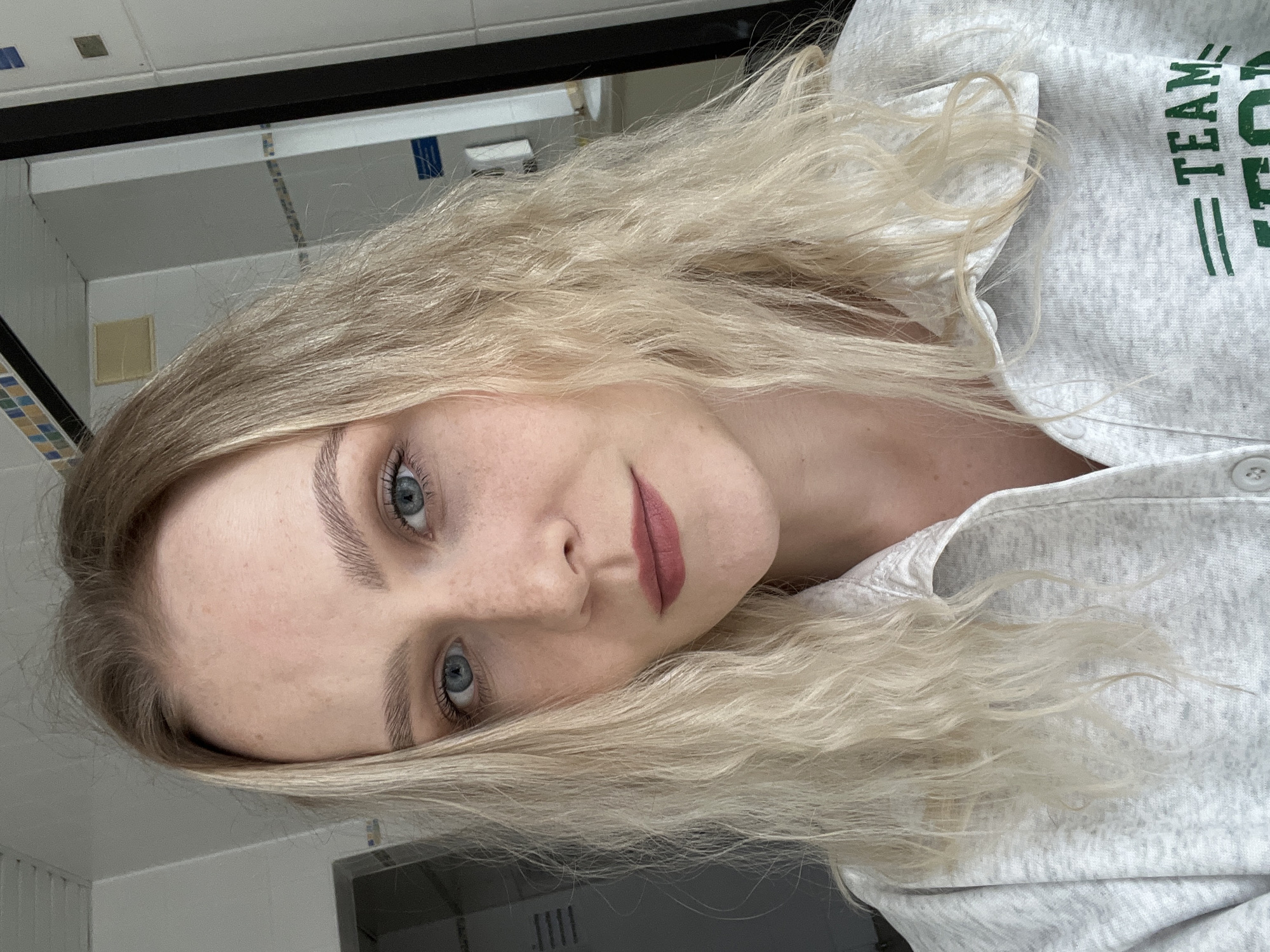 A young Caucasian woman with blonde hair and blue eyes, wearing a grey collared shirt, poses for a selfie in what appears to be a bathroom. She has light makeup on, including lipstick. The background shows tiled walls, a mirror, and a ventilation grate. The scene does not contain any recognizable landmarks or elements to indicate the city of Khimki, Russia.