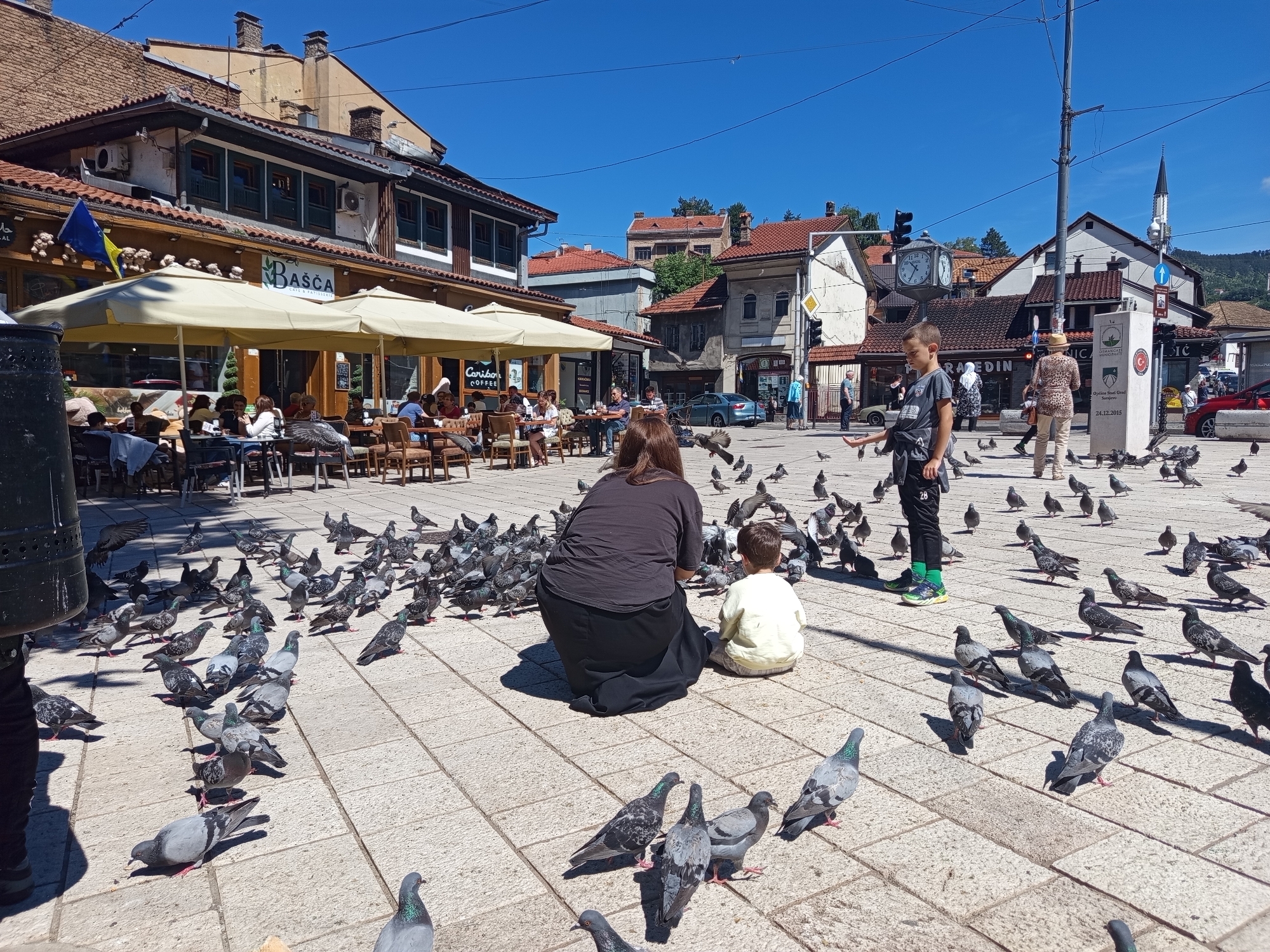 The image depicts a sunlit day in a public square in Sarajevo, Bosnia and Herzegovina, likely Baščaršija, the old bazaar. In the foreground, a large flock of pigeons congregates on the paved square. A woman in a black outfit is kneeling, facing away from the camera, and a young boy in a light-colored shirt sits beside her, also facing away. The boy is reaching out his hand towards the pigeons. Behind them, several other people are scattered throughout the square, some seated at outdoor tables of a cafe with yellow umbrellas. The buildings surrounding the square feature red tile roofs and traditional architecture, with some displaying historical signage and flags, including the flag of Bosnia and Herzegovina. A clock tower is visible in the background, along with the minaret of a mosque. The scene is active, with people enjoying the outdoors and interacting with the pigeons.