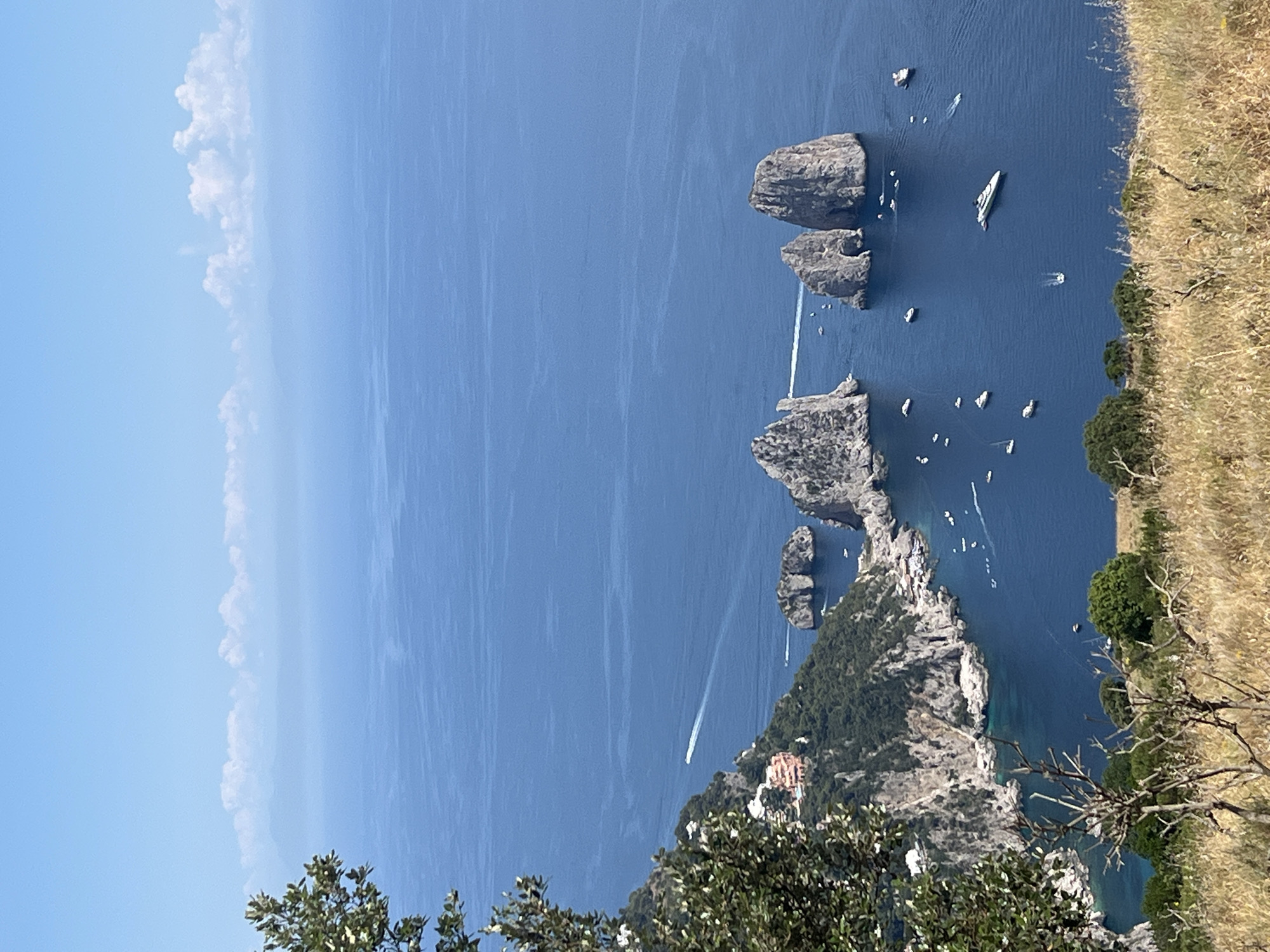 This is an aerial, high-angle, medium shot of the Faraglioni rock formations in Capri, Italy, as viewed from the island's higher elevations. The scene is dominated by the deep blue sea and a bright, partly cloudy sky.
In the foreground, a dry, grassy hillside with scattered twigs and branches frames the lower portion of the image. To the left, a portion of a green, leafy tree juts into the frame.
The middle ground features a dramatic coastal landscape with steep, green-covered cliffs. White buildings are nestled into the side of one of these cliffs. A small, dark rock formation rises from the sea near the coast. Several boats of various sizes, including yachts and smaller vessels, dot the water around the coastline and the iconic Faraglioni. A white speedboat leaves a visible wake as it travels across the water.
The Faraglioni are the prominent feature in the mid-to-far ground, with three large, weathered rock stacks rising majestically from the sea. The largest two are close together, while a third, smaller stack is positioned to their left.
The background consists of an expansive, calm blue sea that stretches to the horizon. In the far distance, hazy mountains are visible, overlaid by a band of soft, white clouds against a pale blue sky. The overall impression is one of a serene, picturesque coastal scene on a sunny day.