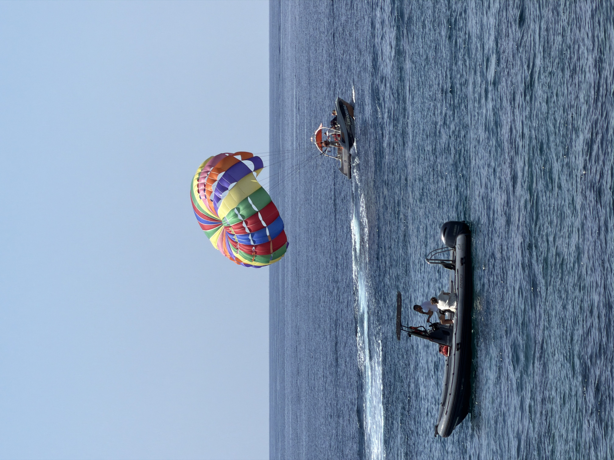 A bright, multi-colored parachute billows in the sky above a blue sea, tethered to a speedboat that is actively moving. Two people are visible on the speedboat, one operating the vehicle and another seated behind. In the foreground, another, larger speedboat with two people on board is also in motion, leaving a wake. The water is a deep blue with moderate waves. The sky is clear and a pale blue. The scene appears to be set on the coast of Budva, Montenegro, a popular tourist destination known for its water activities.
