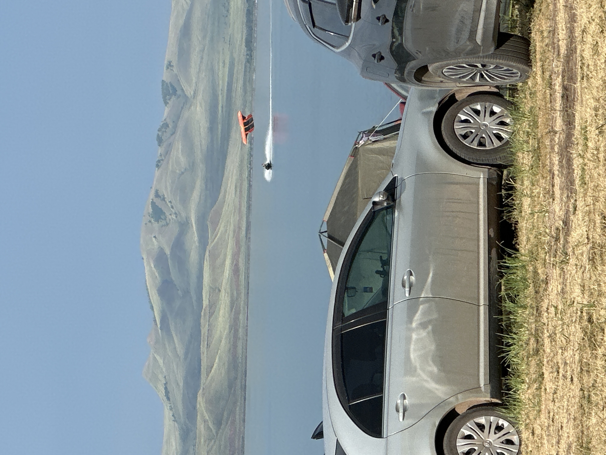 The image shows a bright, sunny day at a large body of water, likely a lake or sea, with rolling hills in the background. Two cars are parked on dry, grassy ground at the edge of the water. One is a dark-colored SUV and the other is a silver sedan. In the distance, a person is waterskiing, leaving a white wake behind them. They are on a bright orange and black watercraft, possibly a jet ski or a towed device, and appear to be in motion. The sky is a clear, light blue, and the overall atmosphere suggests a recreational outdoor activity. The location is identified as Жемчужный, Russia. No visible text is present in the image.