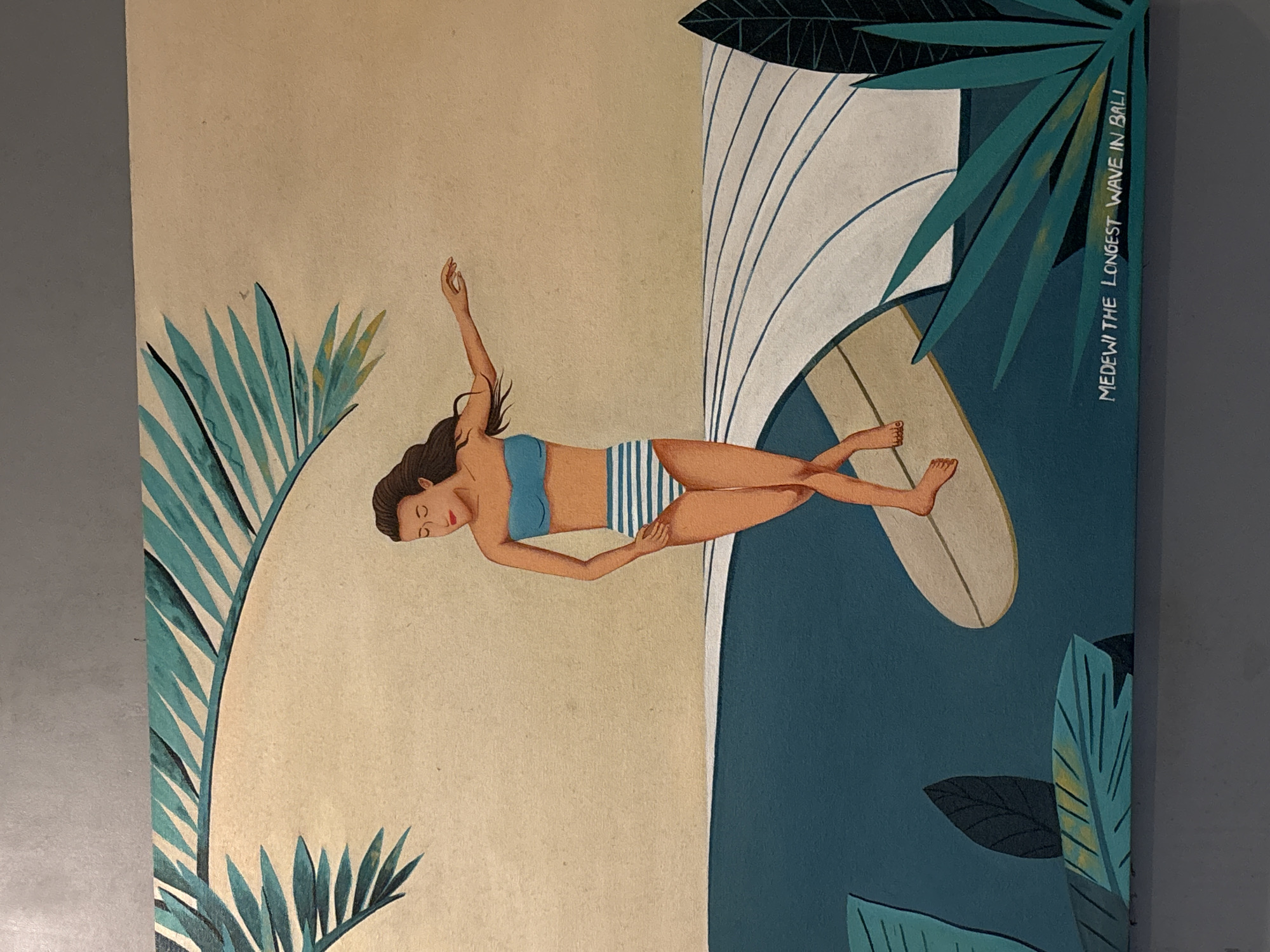 The image depicts a painting featuring a woman lying on a surfboard in what appears to be a stylized depiction of a wave and tropical foliage. The woman is wearing a blue bikini top and striped bottoms. Her hair is brown. She is lying face up with one arm outstretched. The surfboard is cream-colored. Behind her is a tall stylized wave depicted with white lines. Surrounding the scene are various tropical leaves in shades of green and blue. Text on the right side of the image reads "MEDEWI THE LONGEST WAVE IN BALI." This indicates the location is likely Medewi Beach in Pekutatan, Indonesia, known for its surfing.