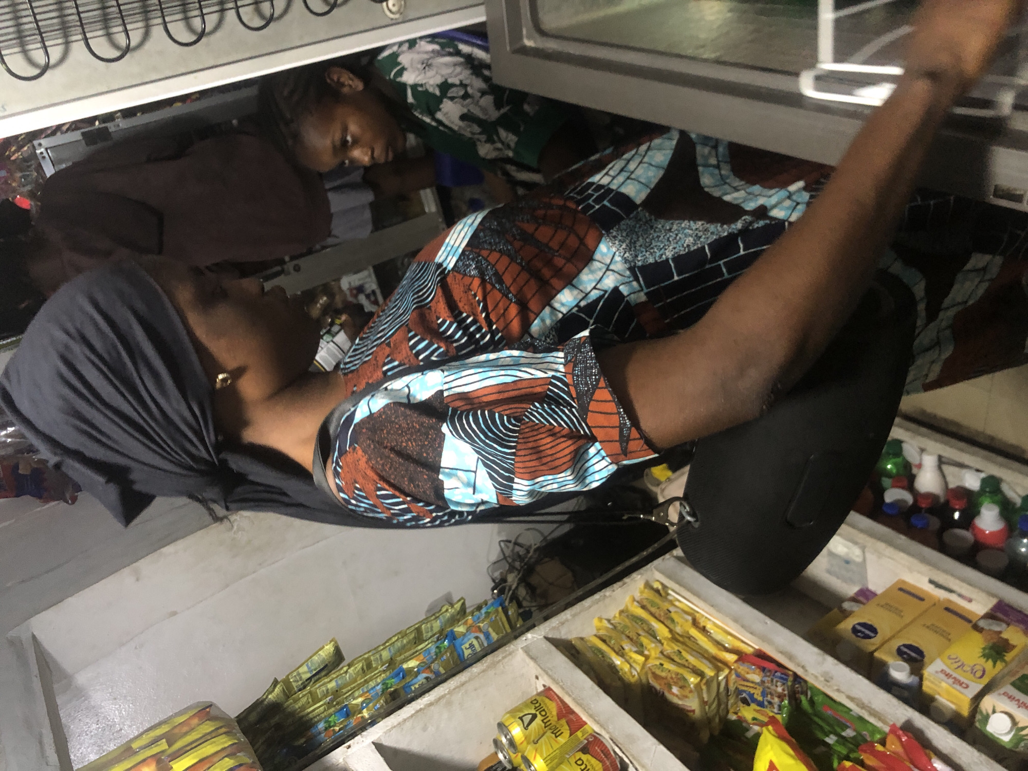 In Ado, Nigeria, a woman wearing a black headwrap and a colorful patterned dress stands in a small shop. She is looking down and to her left. Her left arm is bent and her hand is not visible. A black bag is slung across her body. A young child with braided hair, wearing a green and white floral shirt, is to her right, looking to her left. The shop is filled with shelves stacked with goods, including what appear to be canned drinks, boxes of juice, and packaged food items. A refrigerator is partially visible to the right of the frame. The lighting is dim, suggesting an indoor, possibly low-light environment.