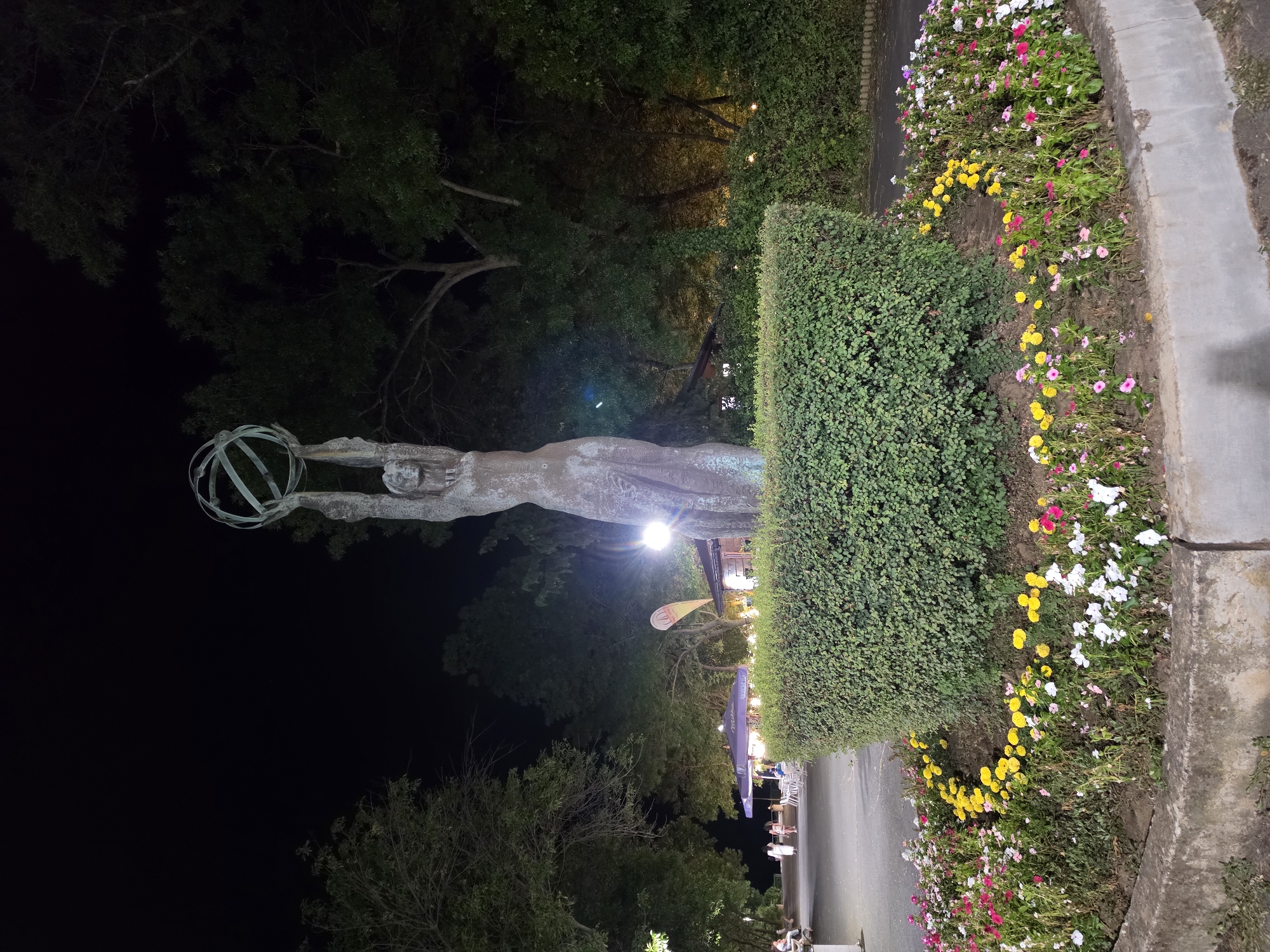 The image shows a night scene in a park in Tsarevo, Bulgaria. In the foreground, a statue of a woman holding a globe aloft stands in the center of a circular flower bed. The flower bed is bordered by a low, trimmed hedge and filled with colorful flowers including pink, yellow, and white blossoms. Behind the statue, a paved walkway leads to an area with outdoor seating, umbrellas, and a building, possibly a cafe or restaurant, illuminated by artificial lights. Several trees with dense foliage are visible in the background, creating a dark, natural backdrop against the night sky. A few people are present in the background, walking along the path or seated at the tables. The overall atmosphere is peaceful and inviting, with the lighting creating a warm glow on the scene.