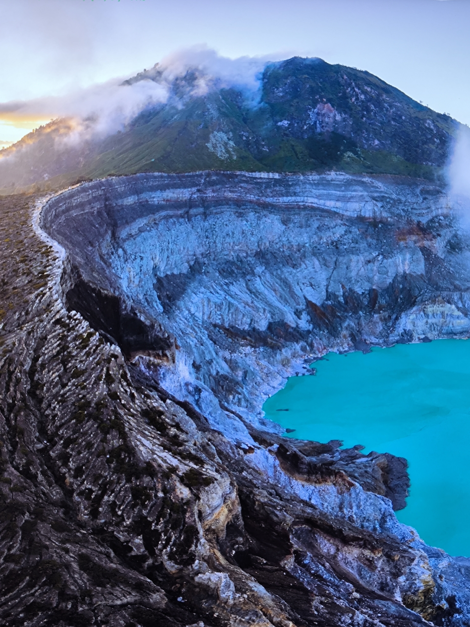 The image displays an elevated view of the Kawah Ijen volcanic crater in East Java, Indonesia, observed under soft, diffused lighting conditions, indicative of either dawn or dusk. The foreground features the rugged, dark grey volcanic rock of the crater rim, characterized by sharp, fractured textures and subtle hints of dark vegetation or moss. A narrow, lighter-colored path or ridge traces the rim's curvature, receding towards the upper left.
The midground is dominated by the steep, stratified inner walls of the crater, which descend sharply to an expansive, opaque, and vivid turquoise-colored acidic lake. The exposed rock faces of the crater walls exhibit distinct layers and varying mineral deposits, presenting a palette of blue, grey, black, and reddish-brown hues. The water in the lake appears uniform in color and texture.
In the background, a large, dark green mountain peak rises, partially enveloped by white, wispy clouds or volcanic gases. The sky above is a gradient from clear light blue to a faint yellowish-orange glow on the far left horizon. No human figures, animals, or man-made structures are visible within the frame. The scene conveys a desolate yet geologically active natural environment.