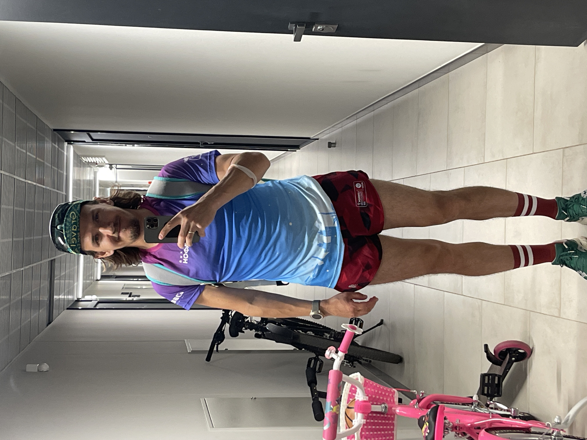 A young adult male is taking a mirror selfie in a hallway. He is wearing a cap, sunglasses, a short-sleeved shirt with a blue and purple gradient, red shorts with a black pattern, red socks with white stripes, and green athletic shoes. He is also wearing a backpack and a watch. A bicycle and a pink children's bicycle are visible in the background. The hallway appears to be in an apartment building or a similar residential complex.