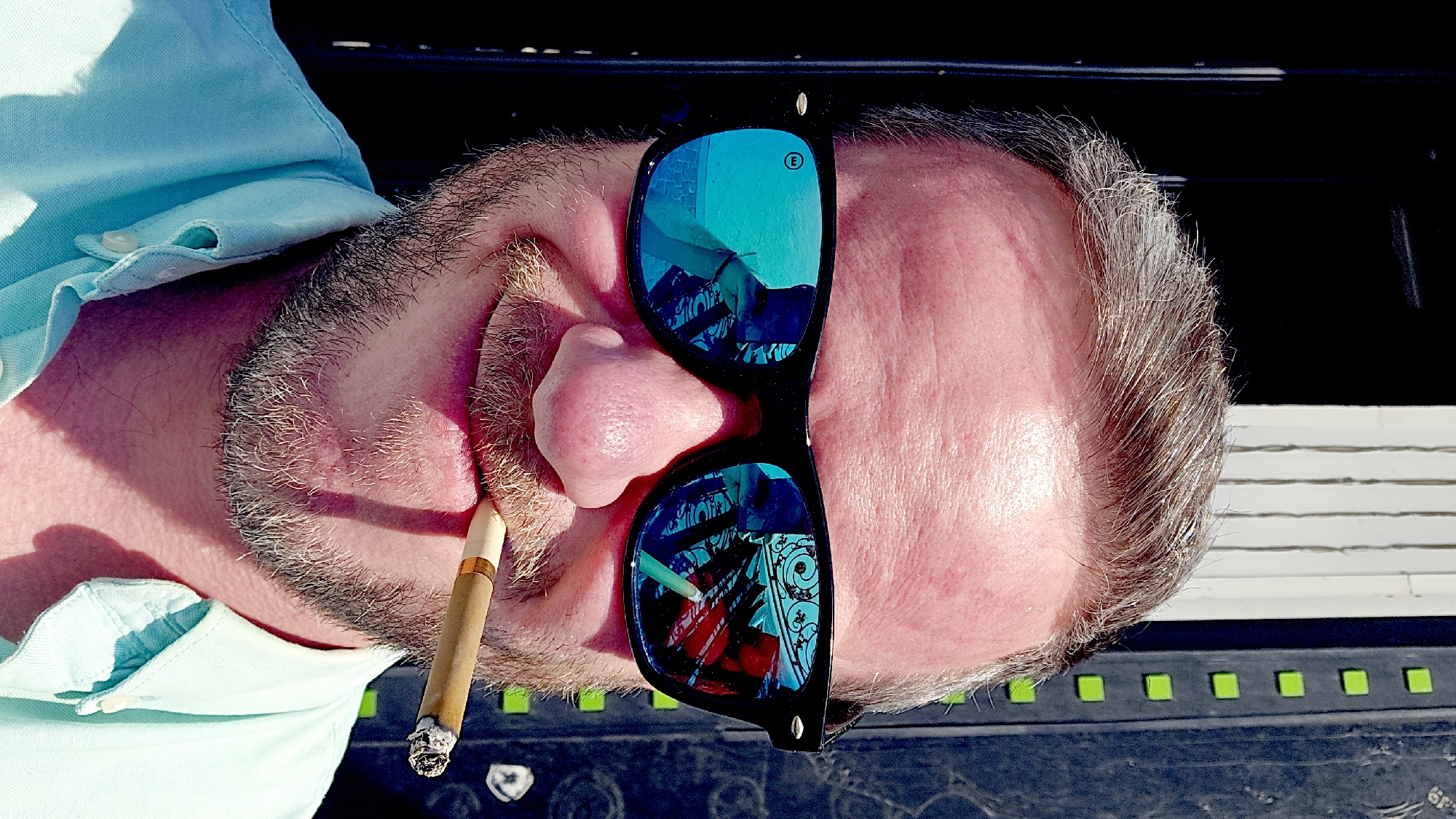 A close-up, slightly angled image shows a man with grey hair and a beard, wearing a light blue shirt and sunglasses. He is holding a lit cigar in his mouth. The sunglasses reflect an urban scene with ornate metalwork, possibly a fence or gate. Based on the reflections, the setting could potentially be Moscow, Russia, known for its elaborate ironwork. The man's face is red, possibly from sun exposure. The background is dark and indistinct but includes a grid-like pattern in black and green.