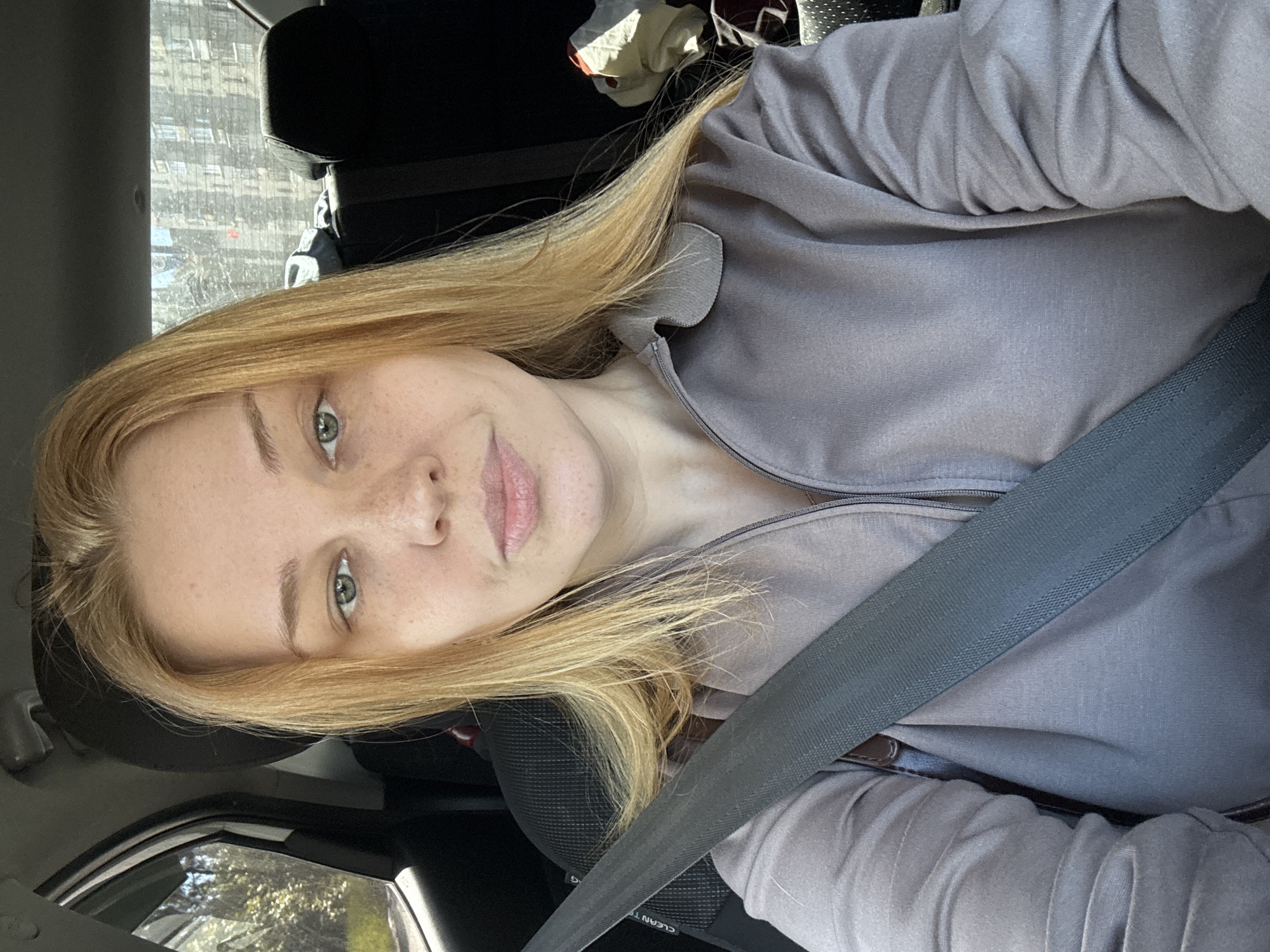 A fair-skinned woman with long, blonde hair and light-colored eyes is captured in a medium shot, rotated to be almost horizontal. She is wearing a light gray or lavender long-sleeved top with a zipper. A dark gray seatbelt crosses her chest and waist. Her facial expression is neutral with a slight hint of a smile. Her eyes are open, and she has visible freckles on her face.
The setting appears to be the interior of a car, indicated by the seatbelt and visible parts of the car's interior, such as what looks like the edge of a headrest behind her and a window to her left. Through the car window, glimpses of what might be buildings or trees are visible, suggesting an urban or suburban environment. The lighting suggests it is daytime, possibly sunny, as some bright areas are visible through the window.
Visible text on the seatbelt is "CLEAN TE".