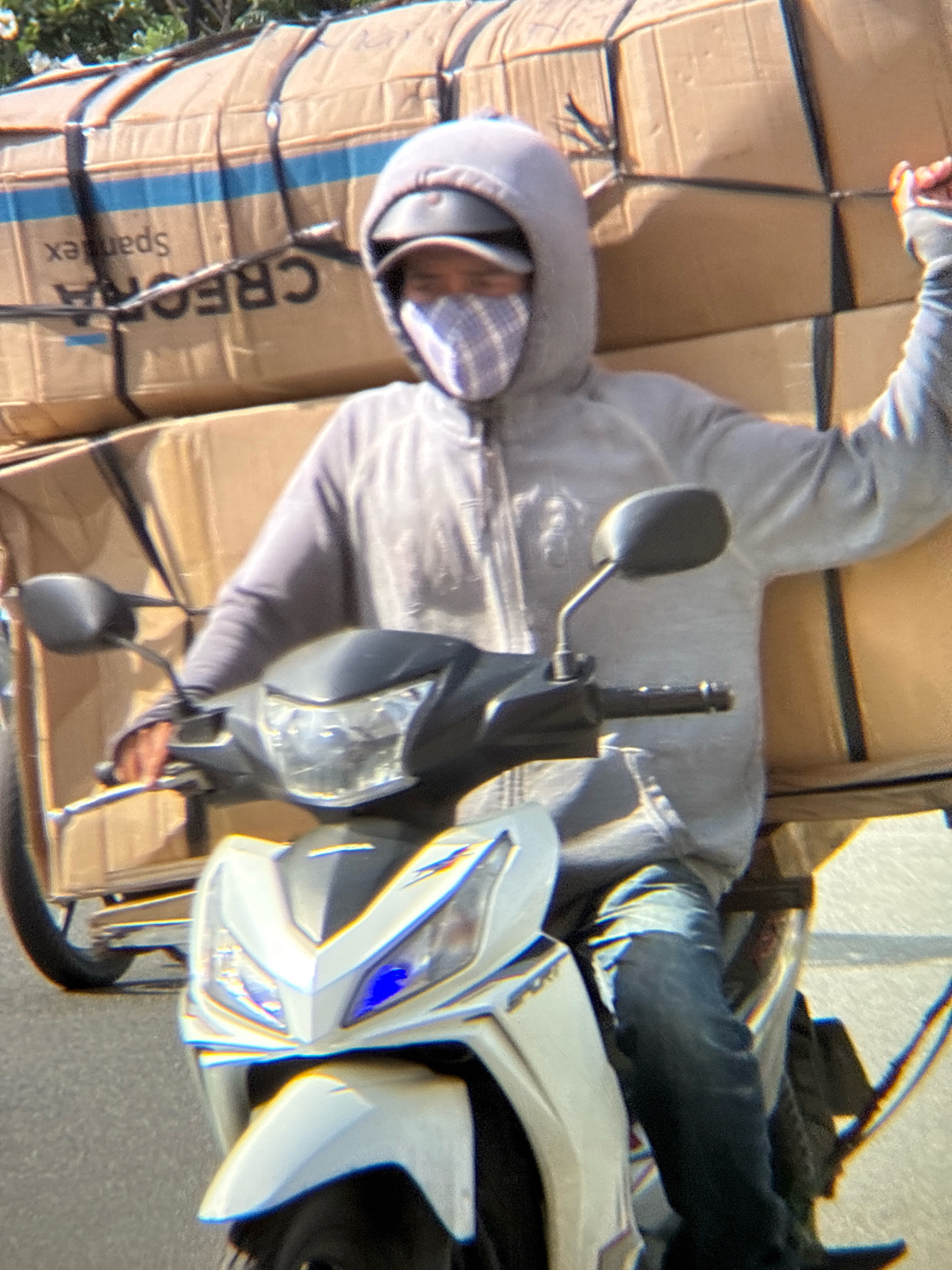 A person wearing a gray hoodie, a checkered mask, and a helmet is riding a white motorcycle through Nha Trang, Vietnam. The motorcycle is loaded with large cardboard boxes stacked high on a trailer. The boxes are strapped together with black straps. One of the boxes has blue and black lettering that reads "CREATION" and "Spantex". The motorcycle has a dark gray front with a blue light. The person is wearing dark blue jeans and has their right arm raised. The scene appears to be on a paved street with some foliage visible in the background.