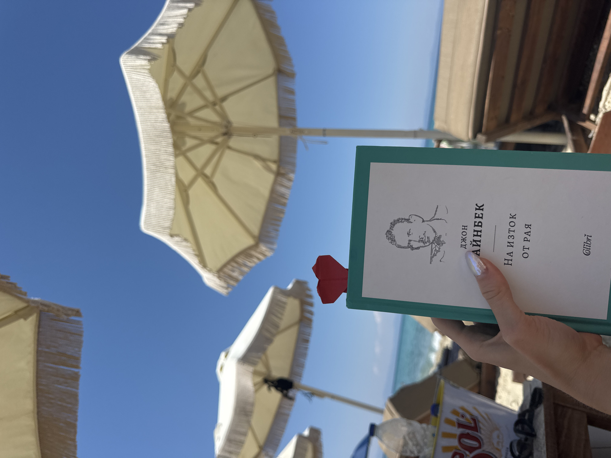 A first-person point of view shot captures a hand holding a teal-covered book with a red origami heart bookmark on a sunny beach in Pallini Municipal Unit, Greece. The book has a portrait of a man and Cyrillic text that reads "ДЖОН АЙНБЕК НА ИСТОК ОТ РАЯ". In the background, two beige beach umbrellas with fringe tassels are partially visible against a clear blue sky. A calm, turquoise sea and a sandy beach with lounge chairs complete the scene. The overall ambiance suggests a relaxing vacation.