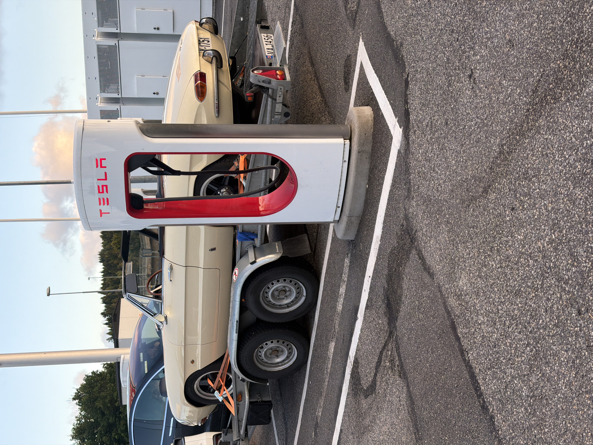 A cream-colored convertible Alfa Romeo Spider is parked on a trailer at a Tesla charging station in Hogstorp, Sweden. The car is facing right, with its front on the left side of the frame. The Tesla charging station is white with red accents and the Tesla logo is visible. The car is being towed by a trailer with two wheels on each side. The trailer is parked in a marked parking spot. The background shows some trees and a building. The sky is partly cloudy. The license plate of the car is V A 1751. The license plate on the trailer is VA 1456.