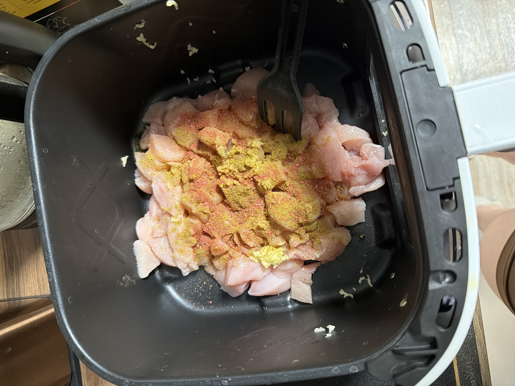 The image shows diced raw chicken in a black air fryer basket. The chicken is topped with a mixture of yellow and reddish-brown spices. A black plastic utensil is partially submerged in the chicken and spice mixture. The basket appears to be part of an air fryer appliance, with a white handle and black trim visible on the right side. Small bits of garlic are scattered around the chicken. The scene is indoors. There is no information to suggest this image was taken in Perm, Russia.