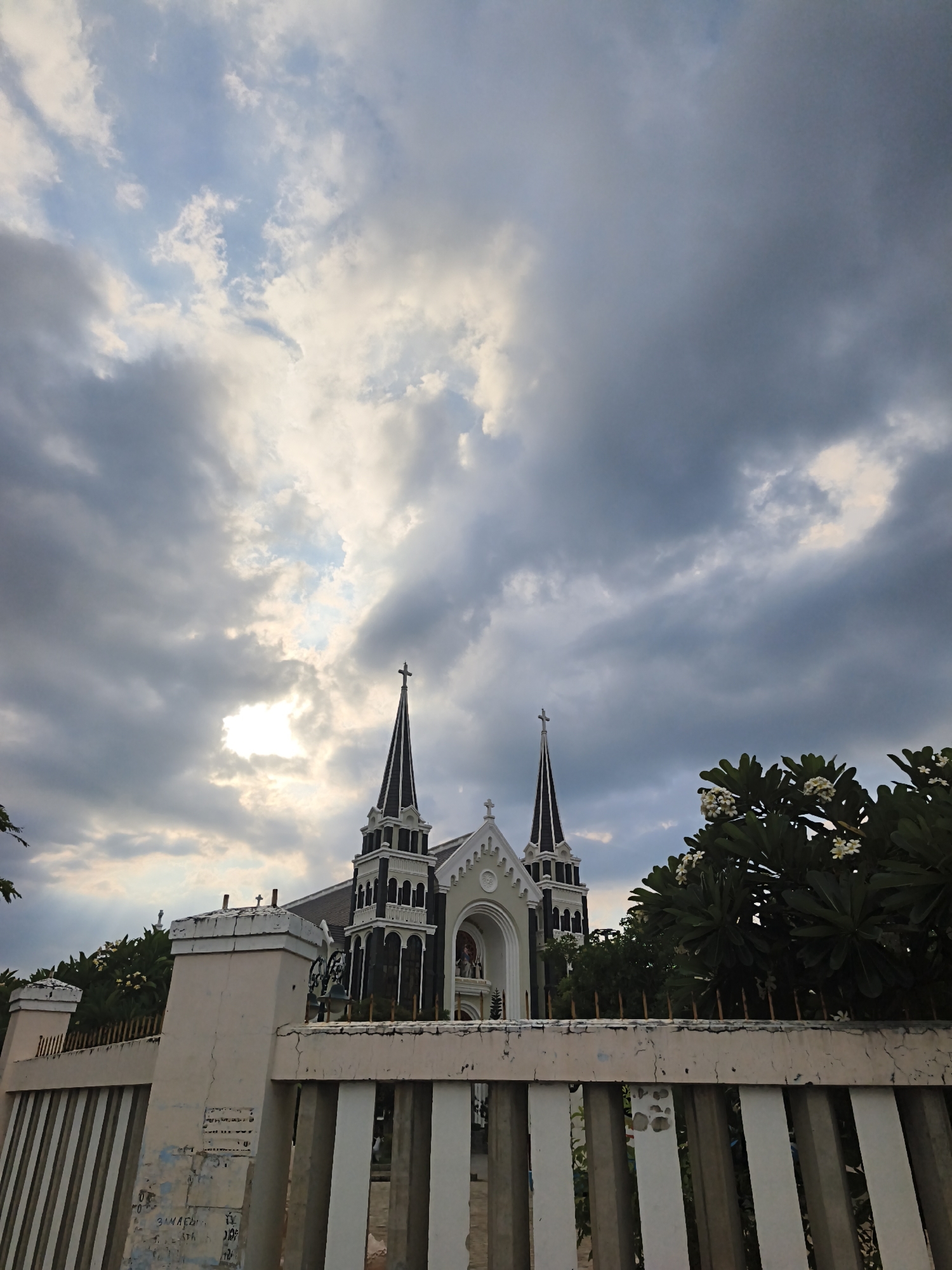 This media file captures a view of a church in Bac Nha Trang Ward, Vietnam, with a dramatic sky overhead. The church, a prominent architectural feature, is Gothic in style with pointed spires topped with crosses. It is painted in dark gray and white, with arched windows and intricate detailing. In the foreground, a weathered white fence with vertical stripes runs across the frame. Beyond the fence, lush green trees with white flowers are visible, framing the church.
The sky is a dynamic mix of dark, heavy clouds and patches of bright sunlight, suggesting either an approaching storm or the aftermath of rain. The light filtering through the clouds creates a dramatic and somewhat moody atmosphere. The time of day appears to be late afternoon or early evening, given the quality of the light.
There is no explicit activity or event taking place; the image seems to be a static shot of the church and its surroundings. The overall emotion evoked is one of solemnity and perhaps a sense of awe due to the imposing architecture and the dramatic sky.
Visible text on the fence includes what appears to be graffiti or faded markings, but they are largely illegible. One section shows what might be Vietnamese characters or numbers.