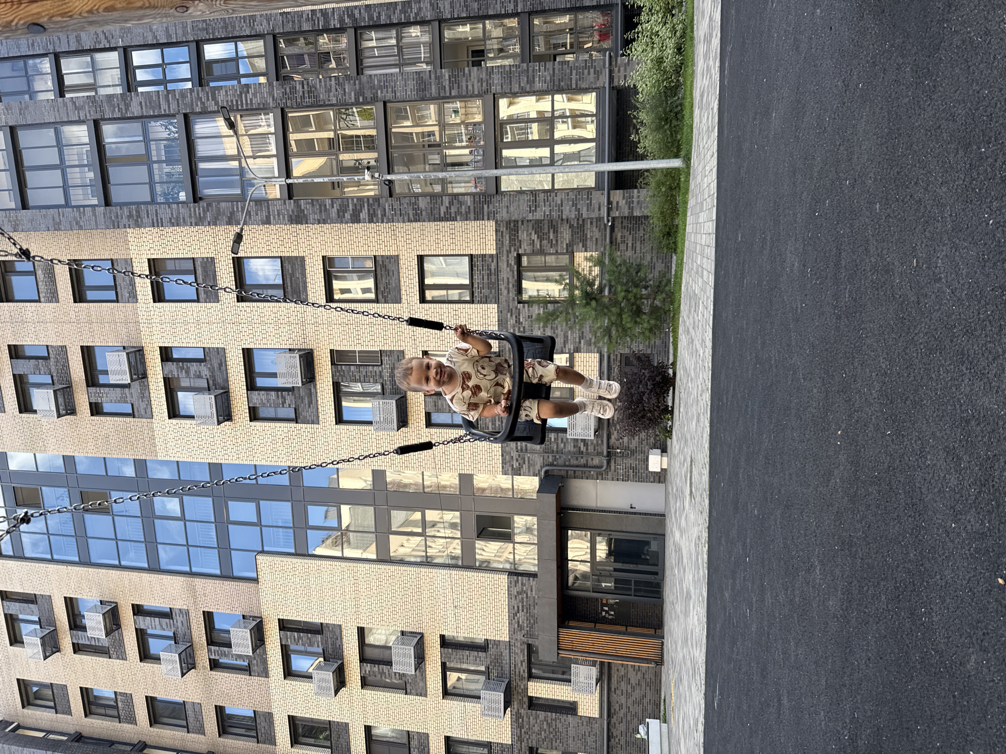 A young child is swinging on a playground swing in front of a modern apartment building. The building has a facade of light-colored brick and dark grey brick, with many windows. The child is wearing a light-colored outfit with a bear pattern and white sneakers. The swing set chains are visible, attached to a frame that is not fully in view. The ground in front of the building is paved with interlocking stones, and beyond that is a dark, soft surface, likely for fall protection on a playground. There are some bushes and a small tree near the building. The overall scene suggests a residential area, possibly a courtyard within the city of Rusanovo, Russia, based on the provided context.