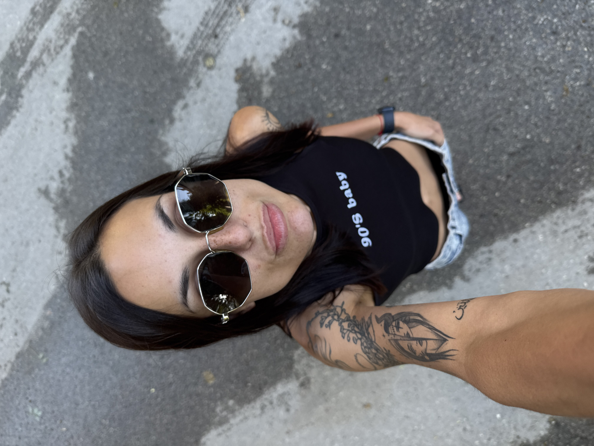 A high-angle medium shot captures a woman with dark hair and sunglasses, looking up at the camera. She is wearing a black crop top that reads "90's baby" and denim shorts. Tattoos are visible on her left arm and shoulder. The woman is standing on a wet, gray asphalt surface, possibly a street or sidewalk in the city of Pleven, Bulgaria. Reflections of trees are visible in her sunglasses.