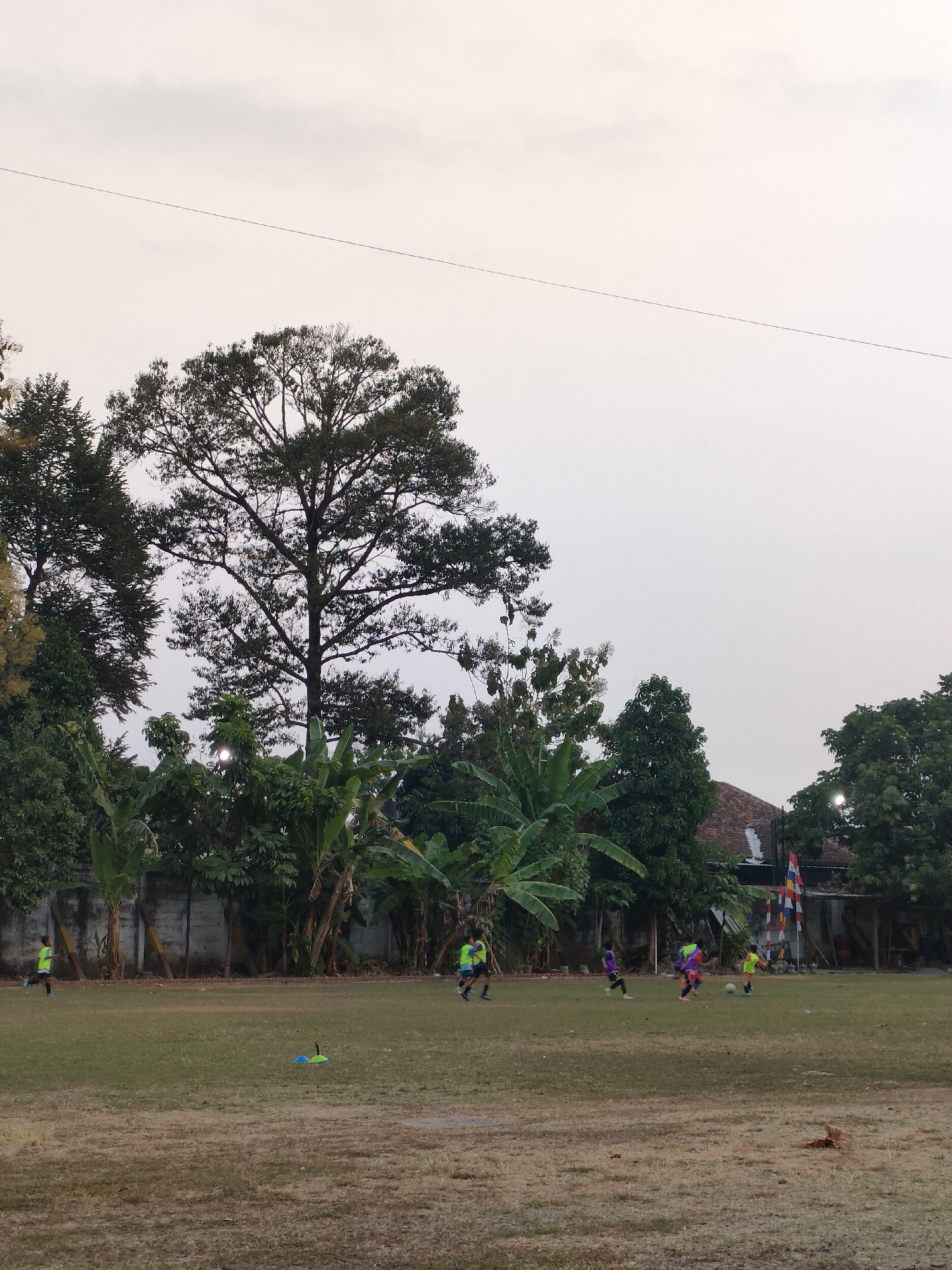 The image depicts an outdoor scene at a grassy field, likely a sports ground, with several individuals engaged in a soccer game. The sky above is overcast with a light gray hue. Dominating the background are lush green trees and foliage, with a large, mature tree with a dense canopy prominently featured on the left side. In the middle ground, there is a low wall, possibly a boundary marker, with several banana trees and other dense vegetation growing behind it. Some buildings with terracotta roofs are visible in the far background, suggesting a semi-urban or rural setting. On the field, several young people, appearing to be children or teenagers, are dressed in athletic wear, predominantly in bright colors such as yellow and purple, and are actively playing soccer. One person is running with the ball, while others are positioned around the field, possibly as teammates or opponents. There are also some cones on the field, likely used for training drills. The overall lighting suggests it might be late afternoon or early evening, given the soft, diffused light. This scene is located in Banguntapan, Indonesia.