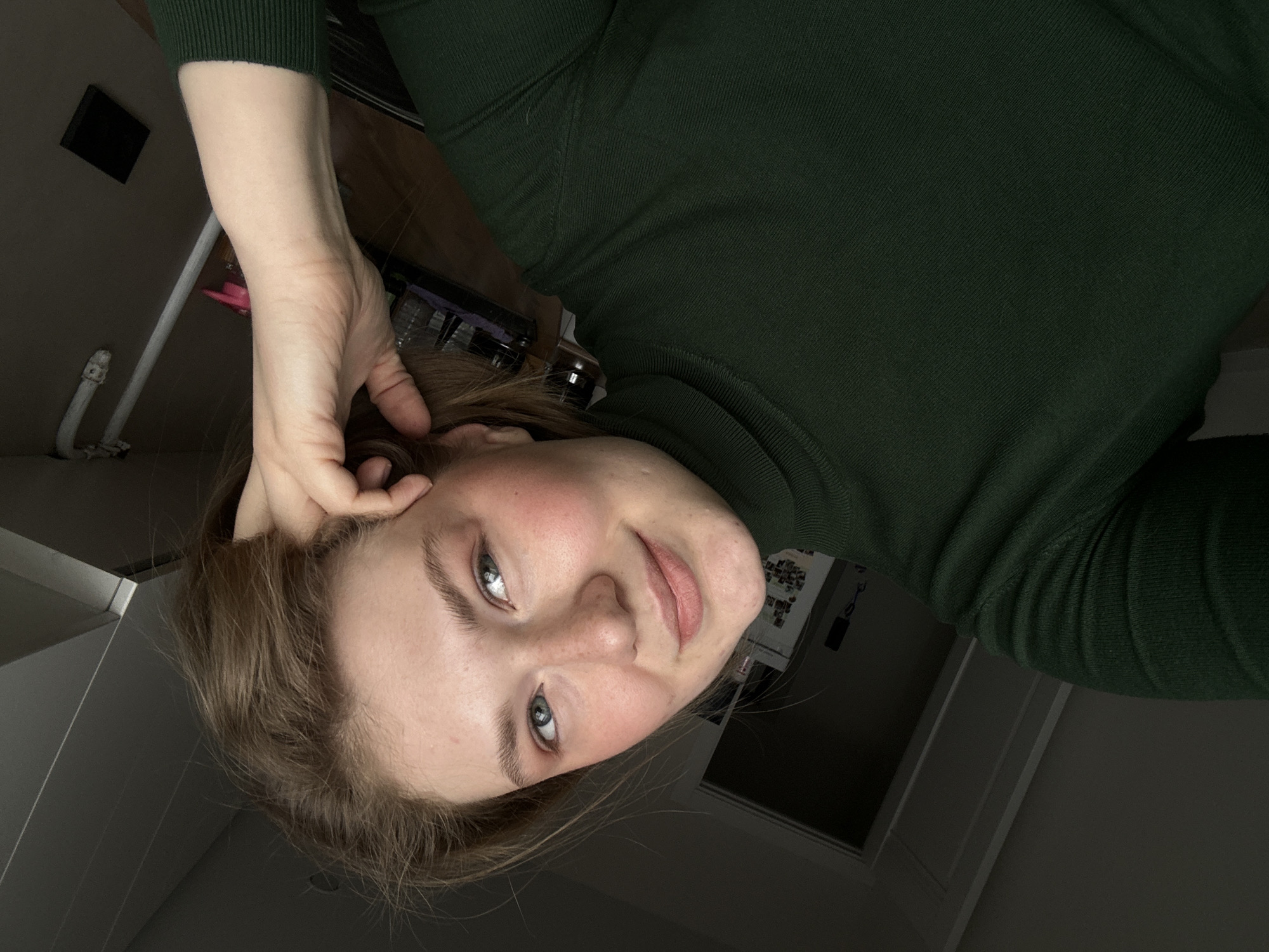 A selfie shows a fair-skinned woman with light brown hair and blue eyes, wearing a dark green turtleneck sweater. She is tilted at an angle, looking into the camera with a slight smile. Her right hand is raised and touches the side of her head near her ear. The background shows a blurred domestic interior with a wooden surface, a wall with pipes, and a door. There is no indication of the city of Kazan, Russia.