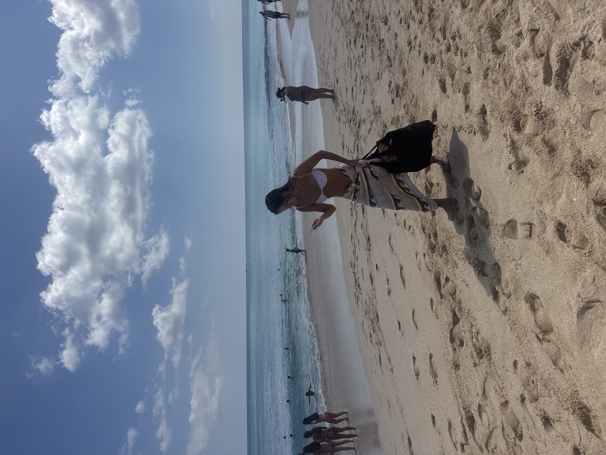 In Kuta Selatan, Indonesia, on a sandy beach, a woman with dark hair, wearing a white bikini top and a patterned sarong, walks towards the viewer while holding a black tote bag in her right hand and a smartphone in her left. She is looking down at the phone. The beach is populated with other individuals; several people are wading or swimming in the shallow turquoise ocean, while a few others are standing or walking on the shore in the background. The sky is blue with scattered white clouds, and the sand is textured with footprints.