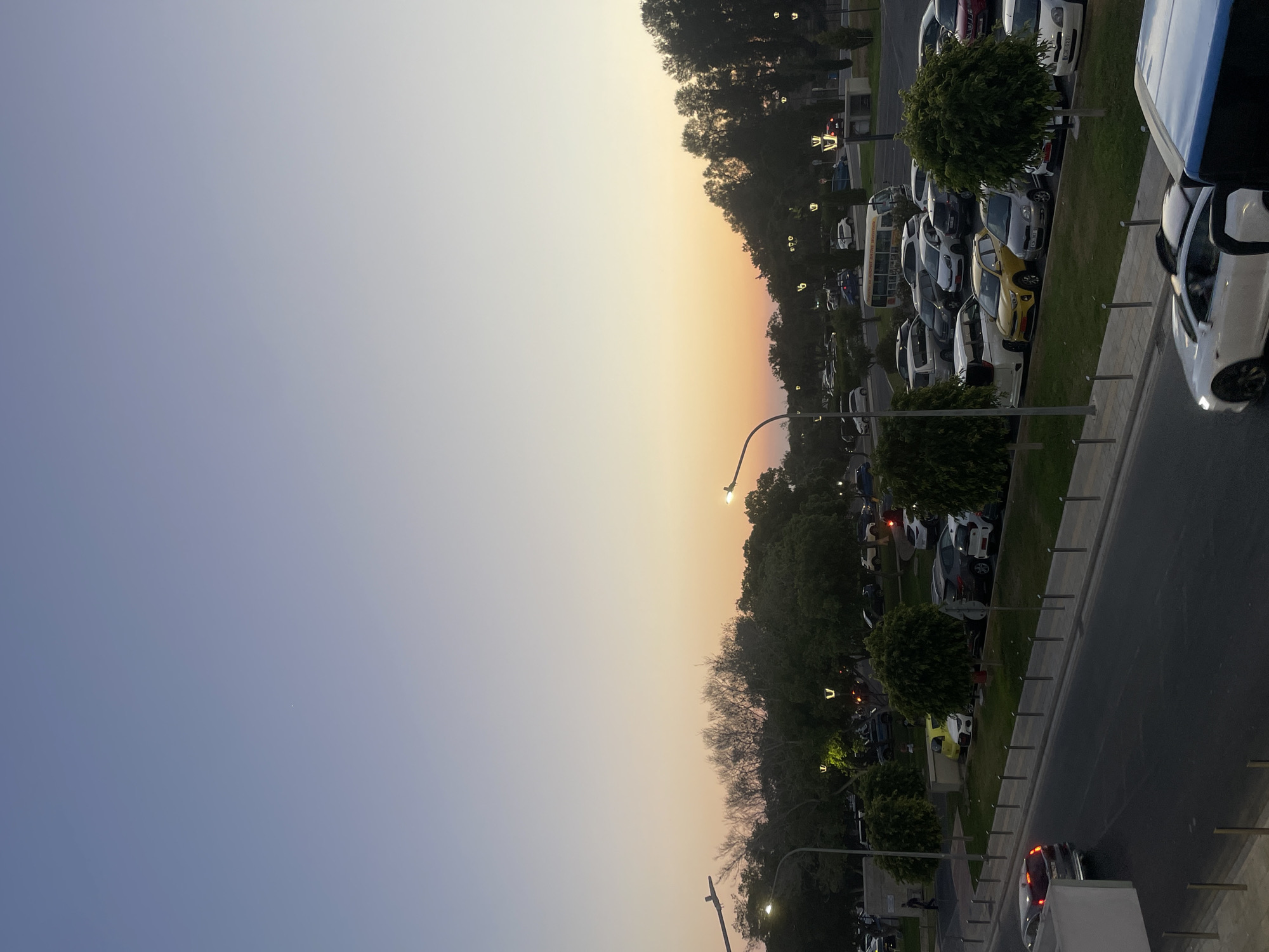 The image depicts a street scene with a parking lot in Paphos Municipality, Cyprus. A clear sky transitions from a pale blue at the top to a soft orange and yellow sunset hue near the horizon. In the foreground, a dark asphalt road curves, with a parked car and a white sports car visible. A blue bus is partially visible on the right side of the image. Beyond the road, a sidewalk is lined with metal bollards, leading to a grassy area with several trees and a parking lot filled with various cars. Streetlights are positioned along the sidewalk and within the parking area. In the background, more trees and buildings are visible. The overall scene captures a quiet evening in a public space.