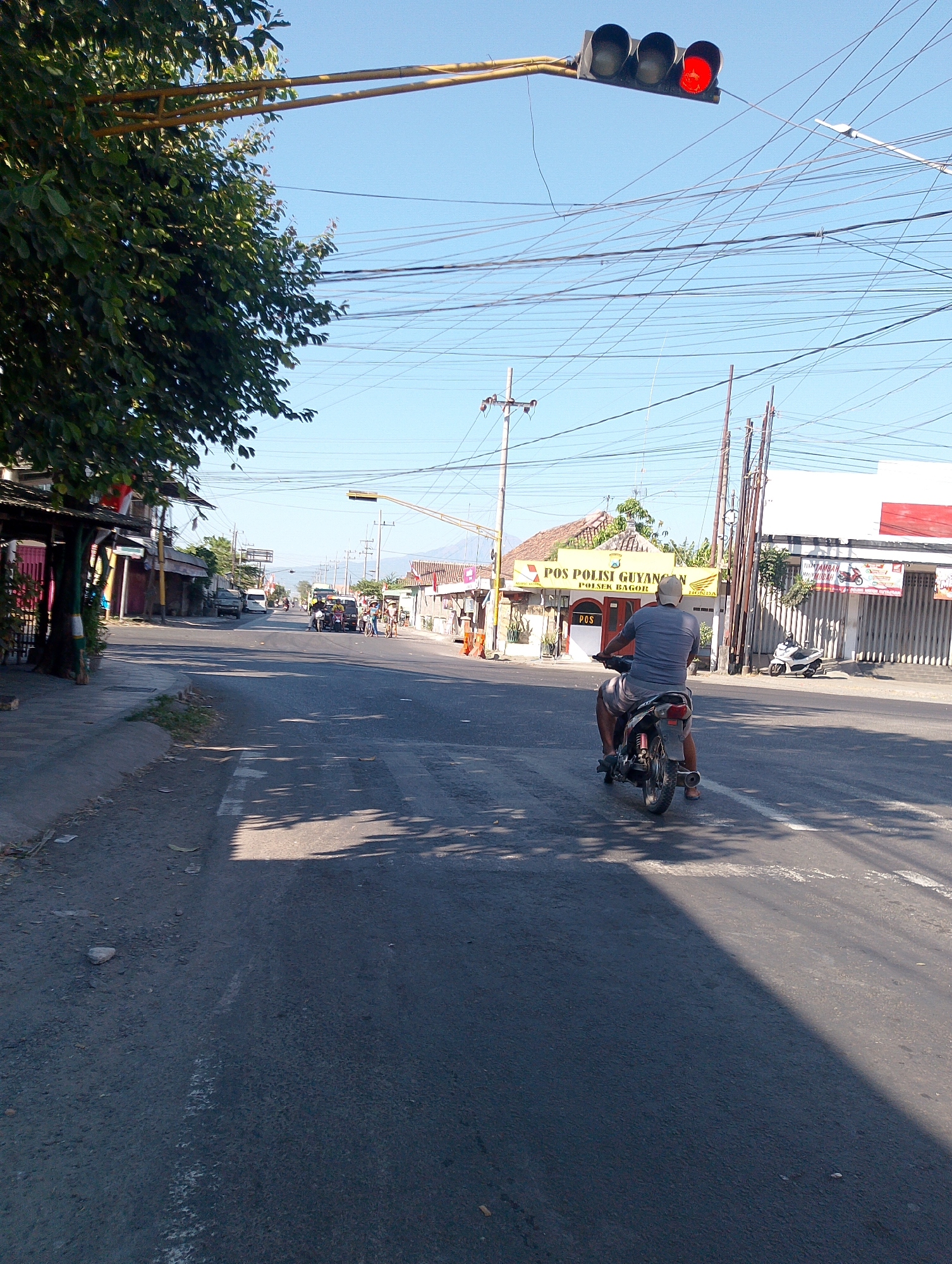 The image depicts a street scene in Guyangan, Indonesia, with a traffic light showing red, indicating that traffic should stop. A man is riding a motorcycle away from the viewer, and other vehicles and people are visible further down the road. The street is lined with buildings and trees, and there are numerous overhead wires crisscrossing the sky. The day appears to be sunny, with shadows cast on the road. The building on the right side of the road has signs that read "TOSAMBA MURAH" and "HONDA". The building on the left side of the road has awnings and appears to be a shop or market. The overall impression is of a typical day in a bustling Indonesian town.
