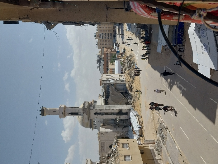 The image captures an elevated perspective of a street in Khan Yunis, Palestinian Territory, under a clear sky with scattered clouds. Dominating the mid-ground is a severely damaged mosque featuring a white minaret with birds perched on its top, and a central dome, both showing visible structural compromise and extensive material loss. Surrounding the mosque and extending into the background are numerous multi-story buildings, many of which are partially collapsed or reduced to rubble piles, indicating widespread destruction across the urban landscape.
A paved asphalt road, marked with white lane lines and a right-turn arrow, traverses the scene from lower-left to mid-right. On this road, approximately five men are walking in various directions, and one man is riding a bicycle towards the viewer. To the right of the road, a larger group of approximately 15-20 individuals, including adults and children, are gathered on an unpaved, debris-strewn area. Further down the street, additional scattered individuals and a dog are visible. In the foreground on the right, the damaged structure of the viewpoint building, exposing concrete and rebar, partially frames the scene. A red-and-white striped object hangs near the right edge, and a black pipe curves across the lower frame. Below, a white tent shelters a seated person, with bicycles parked nearby.