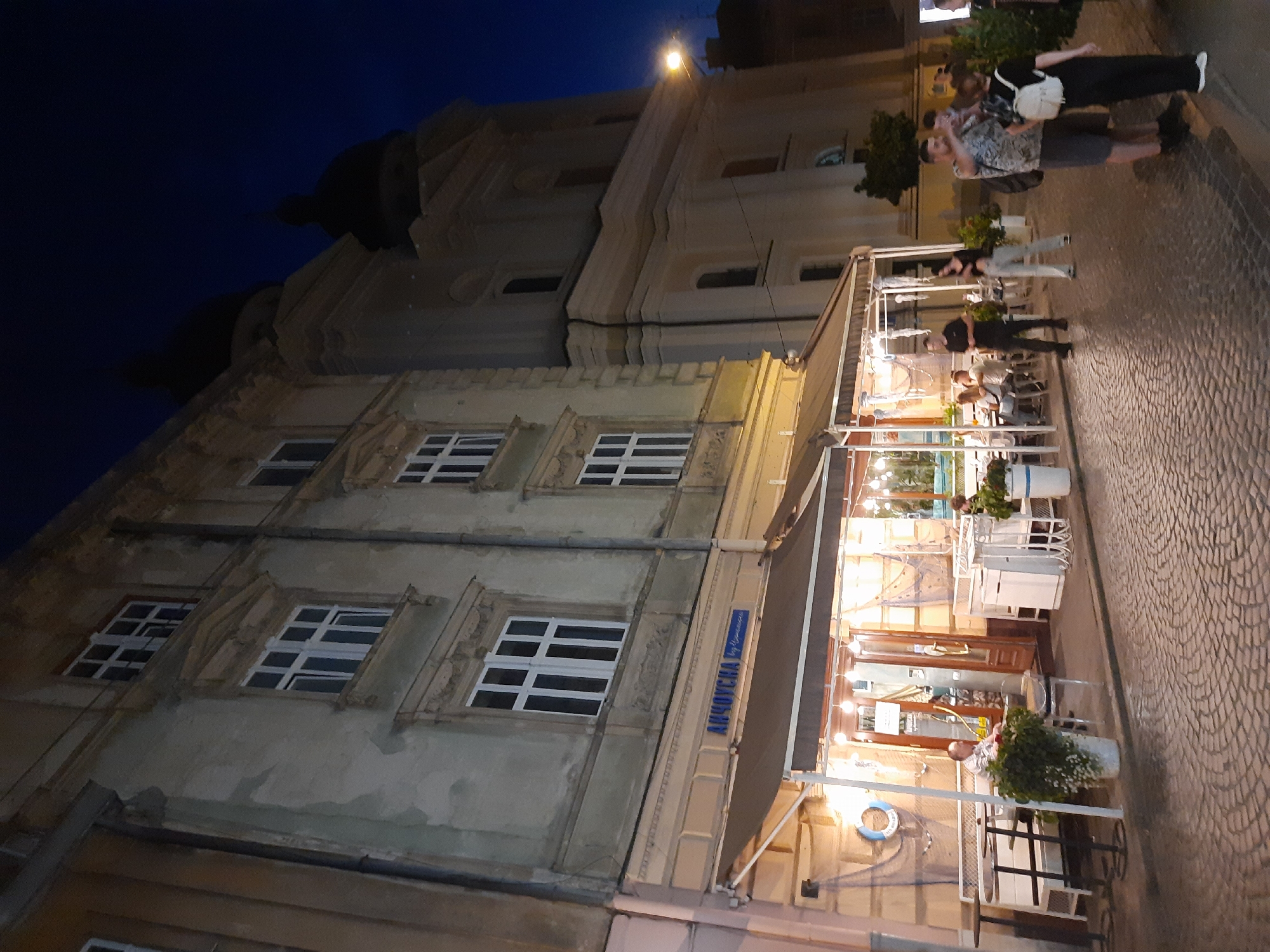 The image shows a nighttime scene on a cobbled street in Lviv, Ukraine, likely in the historic city center. In the foreground, a street-level cafe with outdoor seating is visible, illuminated by warm artificial lights. People are seated at tables, and some are standing outside. The cafe has a brown awning and is decorated with nautical-themed elements, including nets and a life preserver. A sign above the awning reads "АНЧОУСНА by Улюкаєв". To the right, a group of people are standing on the street, some looking at their phones. Further back, a large, light-colored building with multiple windows and architectural details stands tall, possibly a historic structure or church given its design and the presence of domed roofs. The sky is dark blue, indicating nighttime. The street is paved with cobblestones.