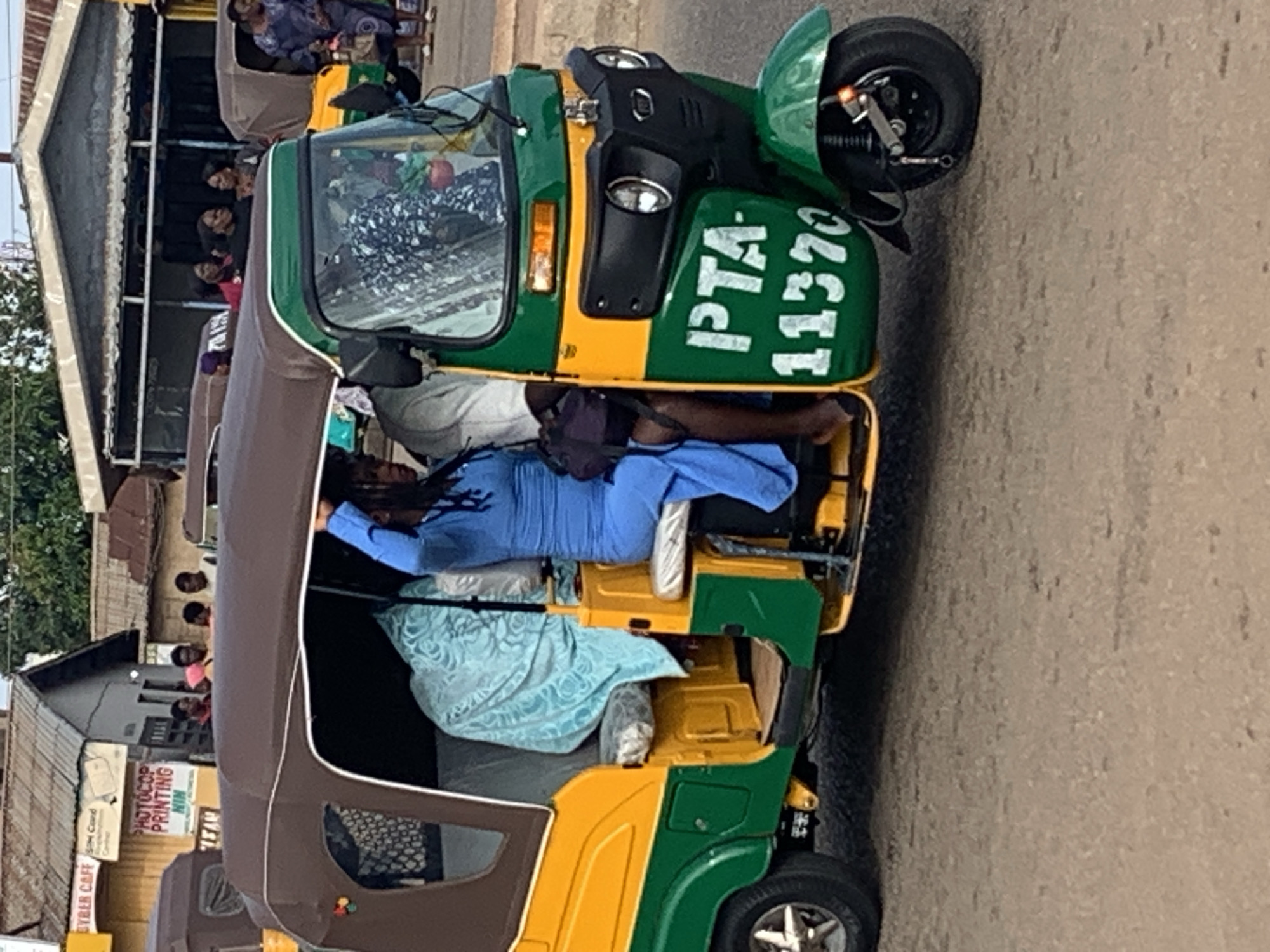 The image shows a street scene in Gura Top, Nigeria. A green and yellow auto-rickshaw, also known as a "Keke Napep" or "Marwa", is parked on the side of the road. The auto-rickshaw has a brown canopy and the registration number "PTA 11370" is painted on its side. A woman wearing a blue outfit is seated in the passenger area of the auto-rickshaw, holding a dark bag. Another person, whose face is obscured, is also visible in the auto-rickshaw. In the background, several other auto-rickshaws are parked, and people are standing near buildings. The buildings have corrugated metal roofs and some signage is visible, including "CYBER CAFE" and "PHOTOCOPY PRINTING". The ground is paved with asphalt. The overall scene depicts a typical roadside environment in a Nigerian city.