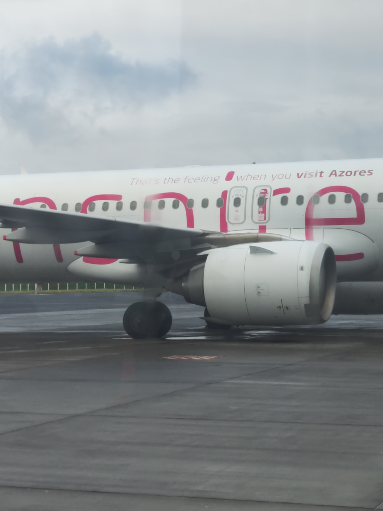 This image depicts the side of a large passenger airplane stationary on an airport tarmac, likely in Ponta Delgada, Portugal. The aircraft's fuselage is predominantly white, adorned with a distinctive pink livery. Part of the airline's slogan, "That's the feeling when you visit Azores," is clearly visible in a smaller, darker font above larger, stylized pink letters that form part of the airline's branding. The letters 'o', 'r', 'e', and 's' from "Azores" are prominent, with the 'e' being particularly large and stylized in pink.
The left wing of the aircraft extends into the frame, and below it, one of the large jet engines is visible. The engine is white with a dark intake, positioned directly above the main landing gear wheel, which rests on the wet tarmac. The ground surface is a dark, wet asphalt or concrete, showing reflections consistent with recent rainfall or wet conditions. The sky above is overcast with a blanket of grey clouds, indicating a cloudy day. No people are visible in the scene. The image captures the plane in a state of rest, possibly awaiting departure or having just arrived at the airport.