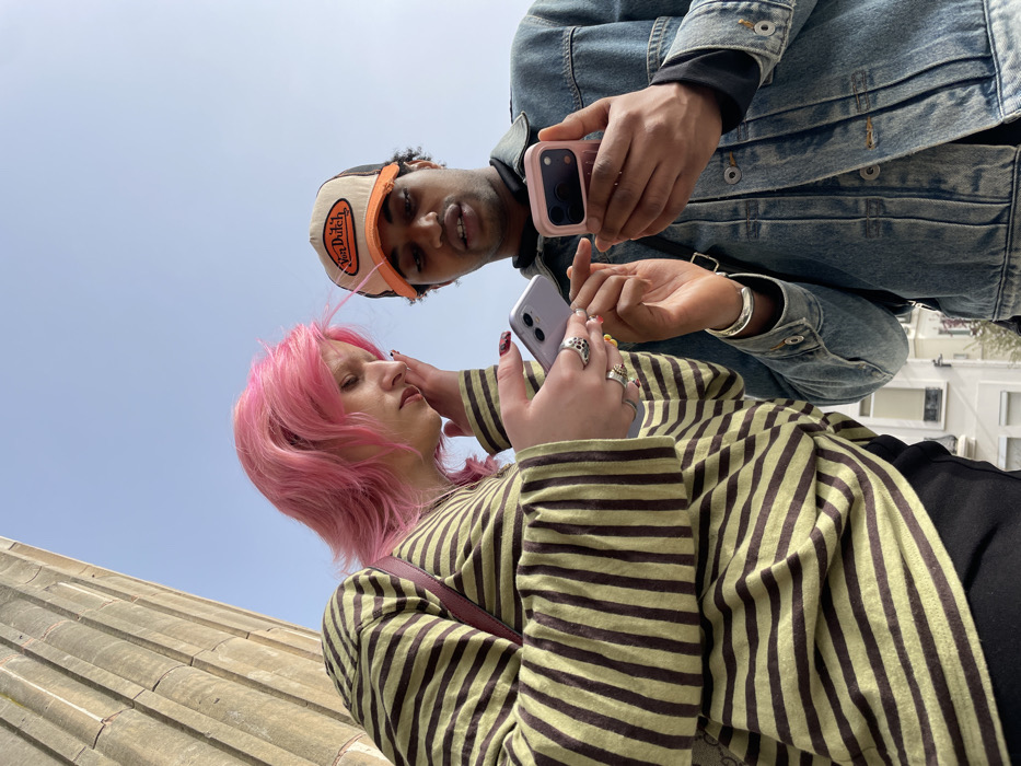 Two individuals are shown from a low-angle perspective, engaged with their smartphones on a clear day in Paris, France, against a backdrop of classical architecture and urban buildings.
The individual on the left has pink hair and light skin, wearing a long-sleeved shirt with alternating light green/yellow and dark brown horizontal stripes, over black pants. A dark strap from a bag or purse is visible over their right shoulder. They wear multiple rings on their fingers, some with prominent designs, and their fingernails are painted in various colors. This individual holds a lavender-colored smartphone in both hands, their head tilted downwards as they focus on the screen. Their left hand is raised towards their ear/hair.
The individual on the right has dark skin and short, dark curly hair visible beneath a distressed tan and orange trucker hat featuring a "Von Dutch" logo. They wear a light blue denim jacket over a dark-colored long-sleeved shirt. A silver cuff bracelet adorns their left wrist, and a black strap from a bag is visible over their left shoulder. This individual holds a pink-cased smartphone in both hands, with their right index finger extended towards the screen, suggesting interaction or pointing. Their mouth is slightly open.
Both individuals are positioned closely, looking at their respective phone screens. The individual on the right appears to be showing or pointing at content on their device.
The immediate background on the left features a large, vertically fluted, light-colored stone column, indicative of a significant classical or neoclassical building, characteristic of Parisian landmarks such as the Pantheon or Église de la Madeleine. The upper background is a clear, light blue sky. In the lower background, behind the individuals, are multi-story, light-colored residential buildings with windows, typical of Parisian architecture.