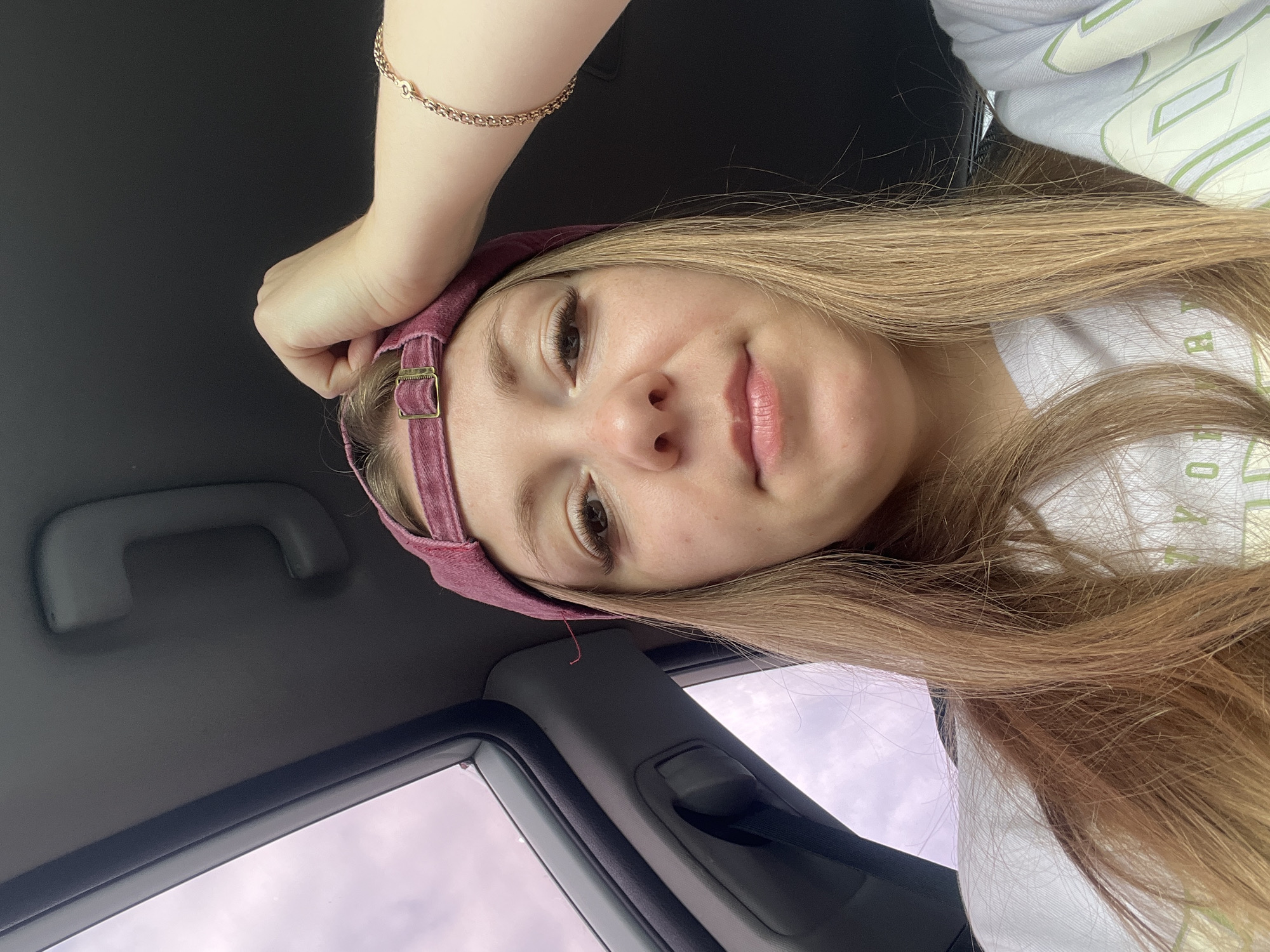 The image shows a young woman in the front passenger seat of a car, looking directly at the camera. She is wearing a maroon baseball cap and a white t-shirt. Her right hand is resting on top of the cap, and she is wearing a gold bracelet on her right wrist. The background shows the interior of the car, including a gray ceiling handle and a sky visible through the open window. There is no visible indication of the city of Shadrinsk, Russia.