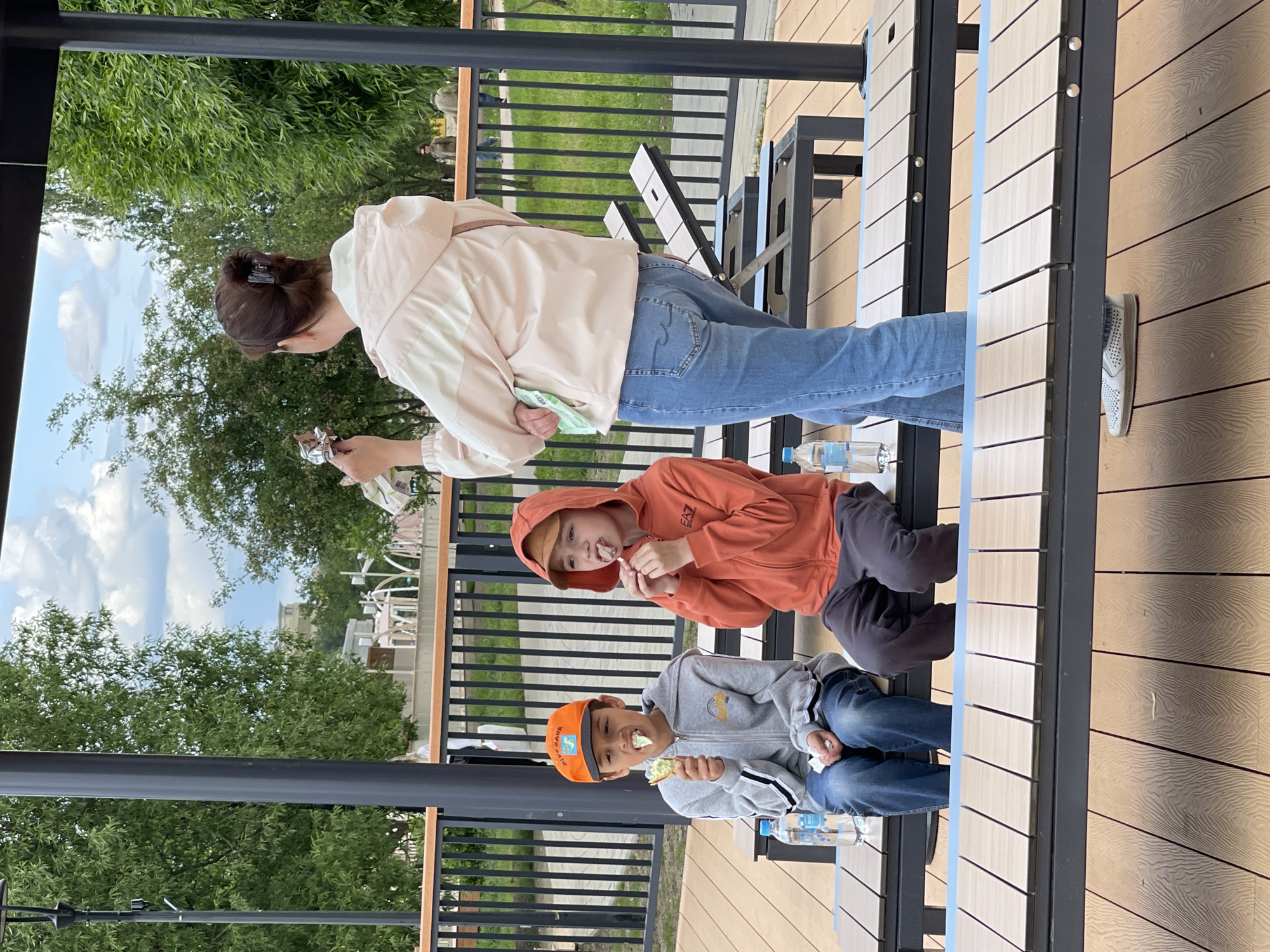 Two boys and a woman are seated at an outdoor table. The two boys are sitting, wearing hooded sweatshirts. One boy is wearing a grey hoodie with blue jeans and an orange baseball cap, and the other boy is wearing an orange hoodie with black pants. The woman is standing, wearing a cream-colored jacket and blue jeans. All three are eating something, likely ice cream. There are water bottles on the table. The table is made of metal and wood, and the ground is made of wood planks. There is a metal railing in the background, with trees and a building visible beyond the railing. The location appears to be Moscow, Russia.